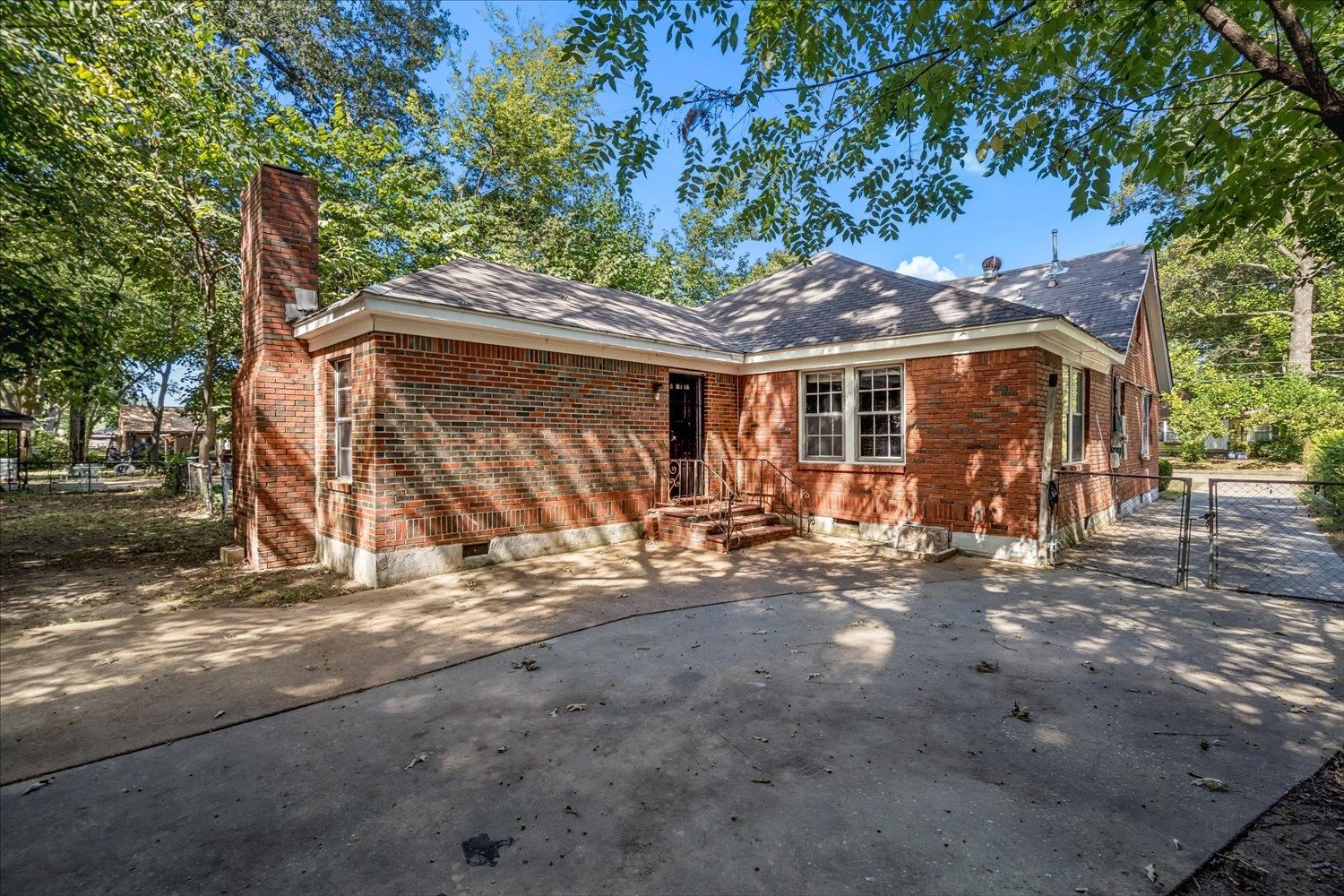 Red brick house with a tall chimney and wide driveway beneath mature trees.