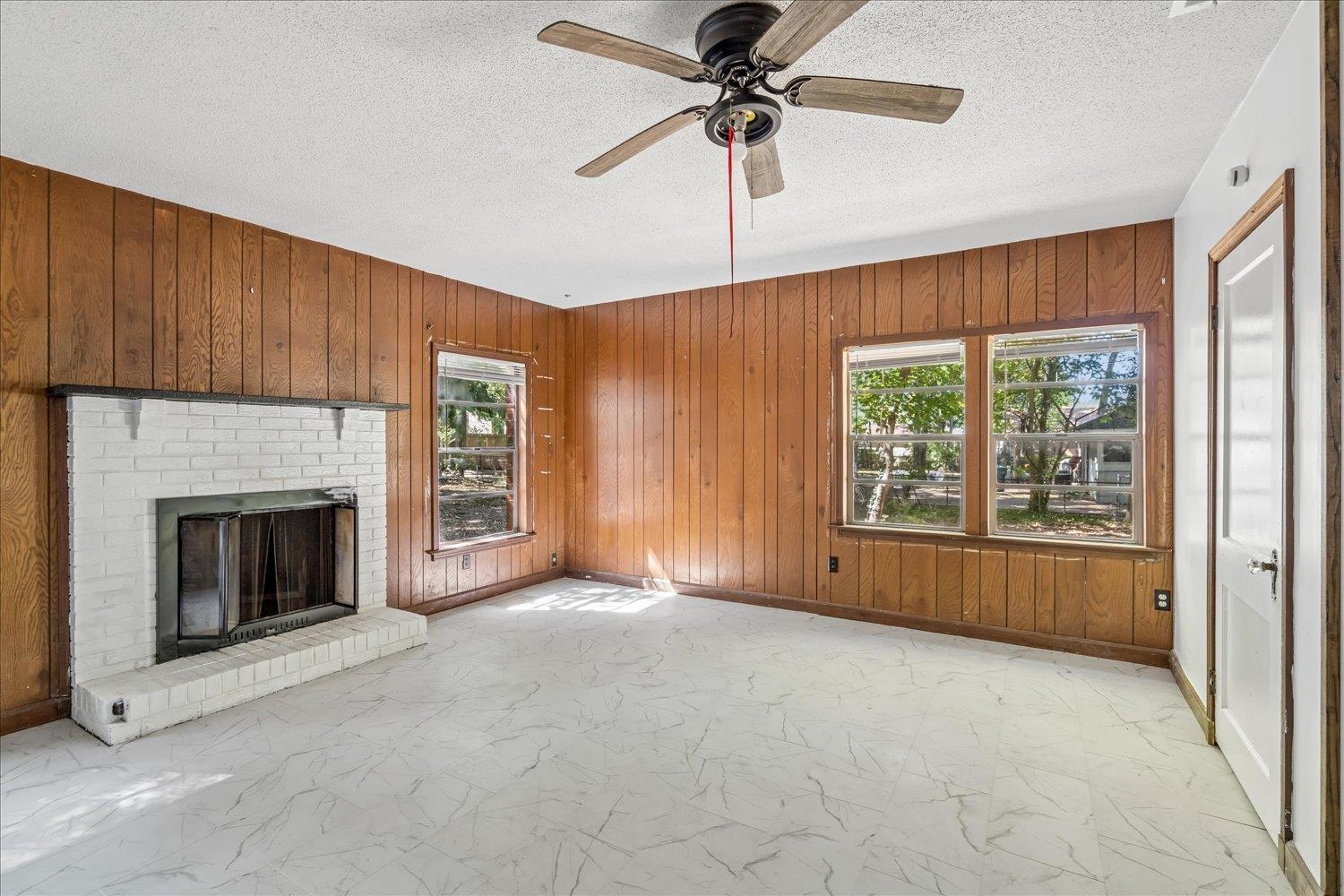 Vacant living room with a white brick fireplace, wood-paneled walls, and a ceiling fan.
