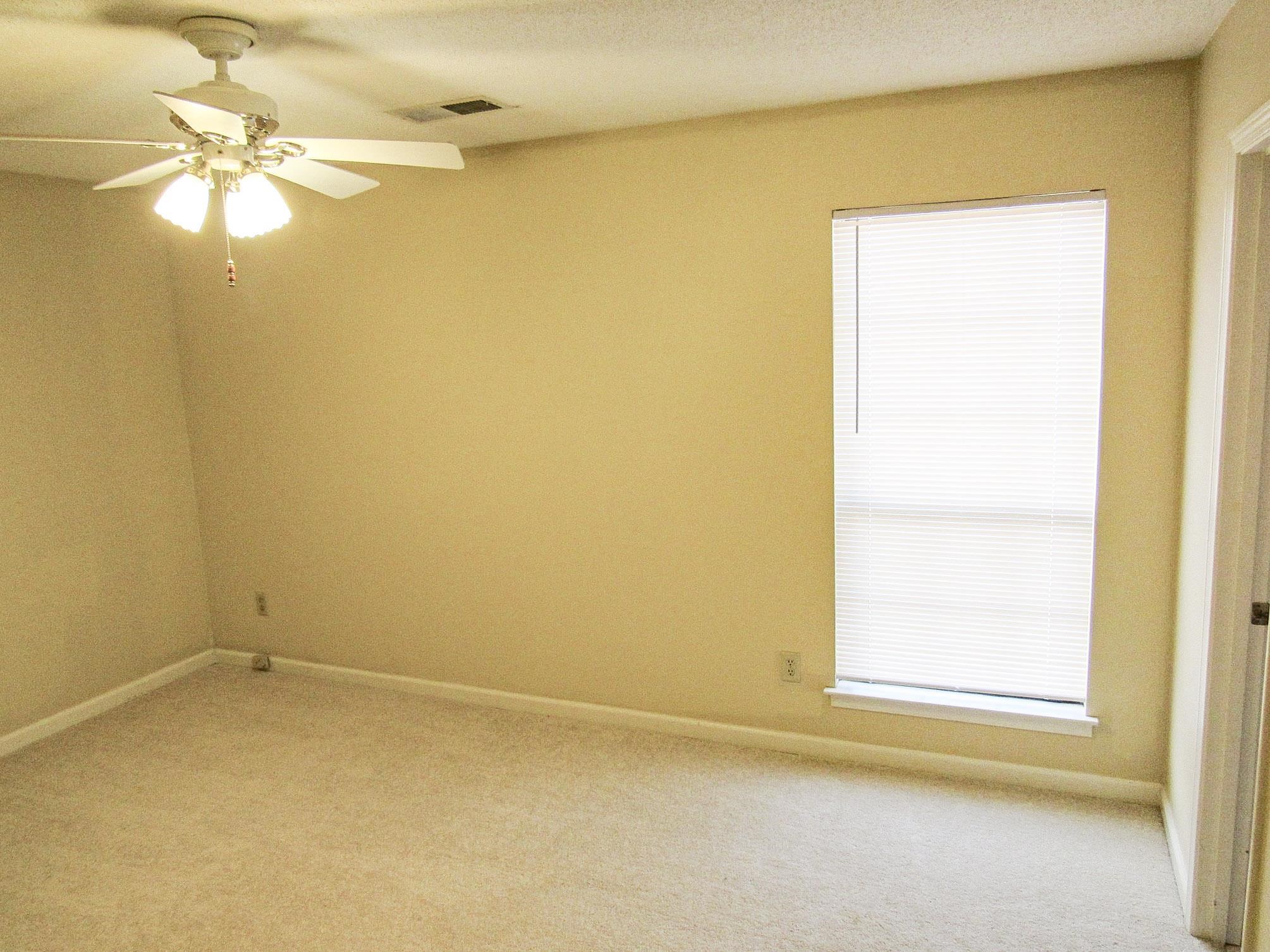 Empty carpeted room with beige walls, a ceiling fan, and a window with white blinds.