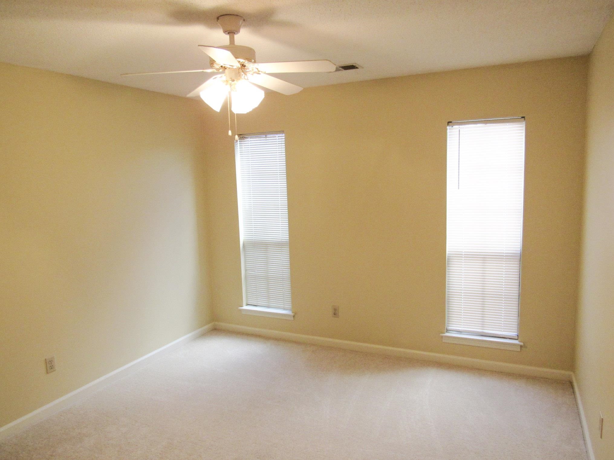 Empty carpeted bedroom with two blinded windows and a ceiling fan with lights.