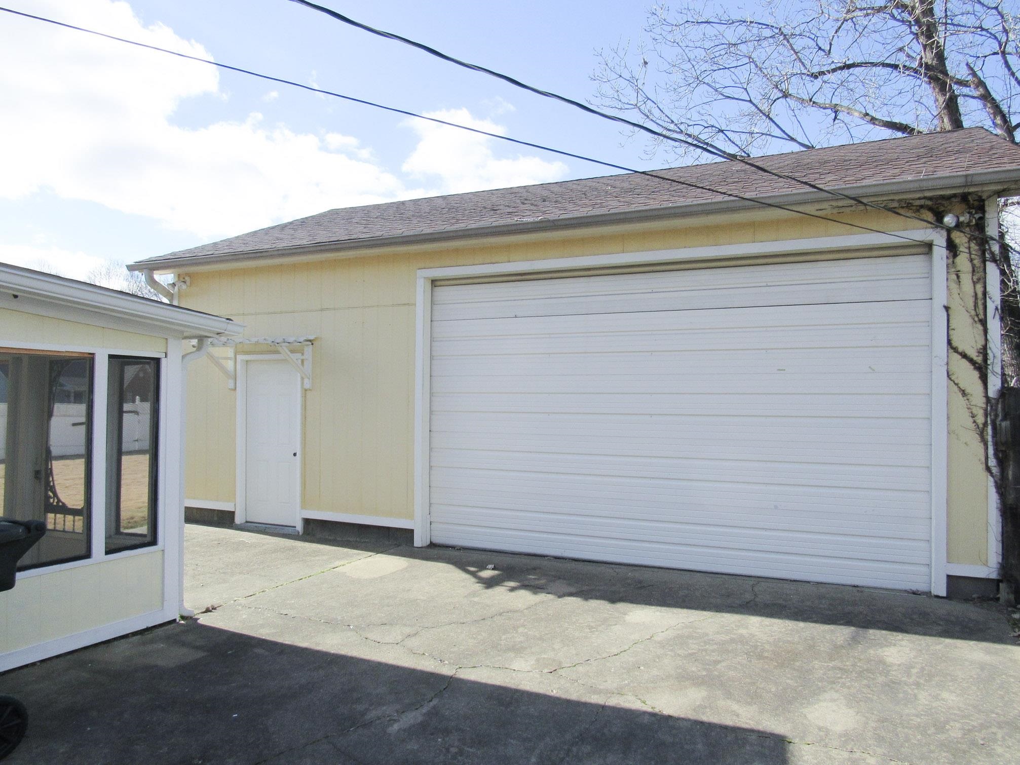 Pale yellow garage with a large white roll-up door beside a concrete driveway.