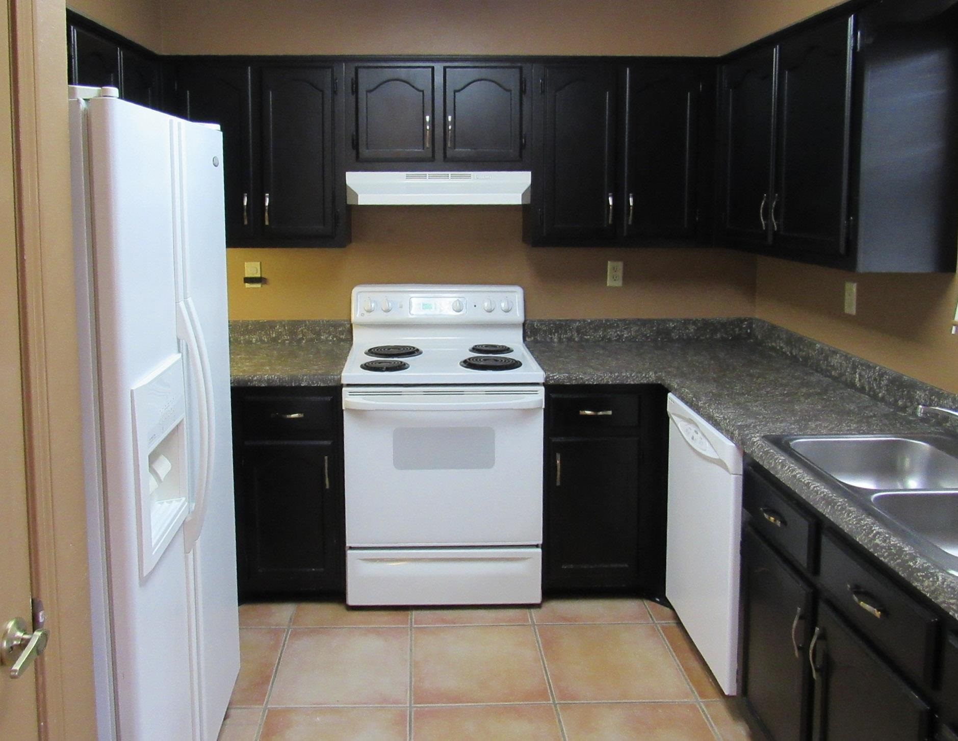 Kitchen with dark cabinets, speckled counters, white appliances, and terracotta-colored tile flooring.