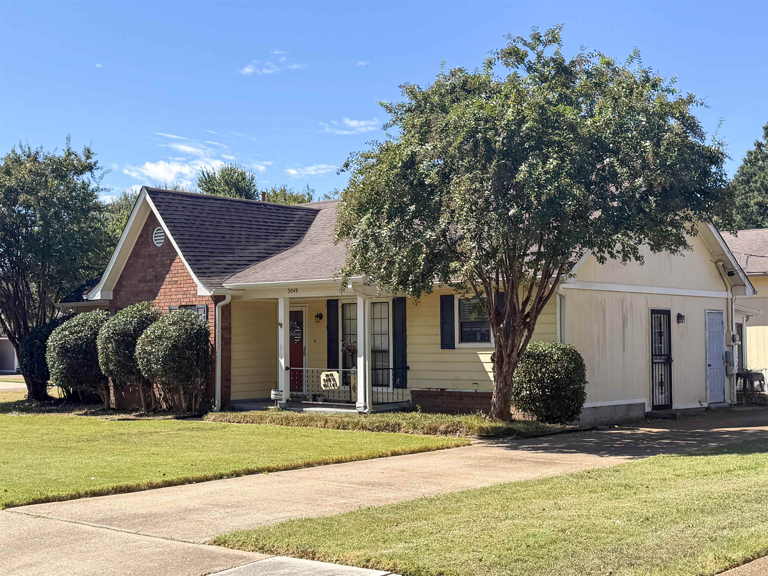 One-story cream-sided home with a red brick gable, covered porch, and large shade tree in front.