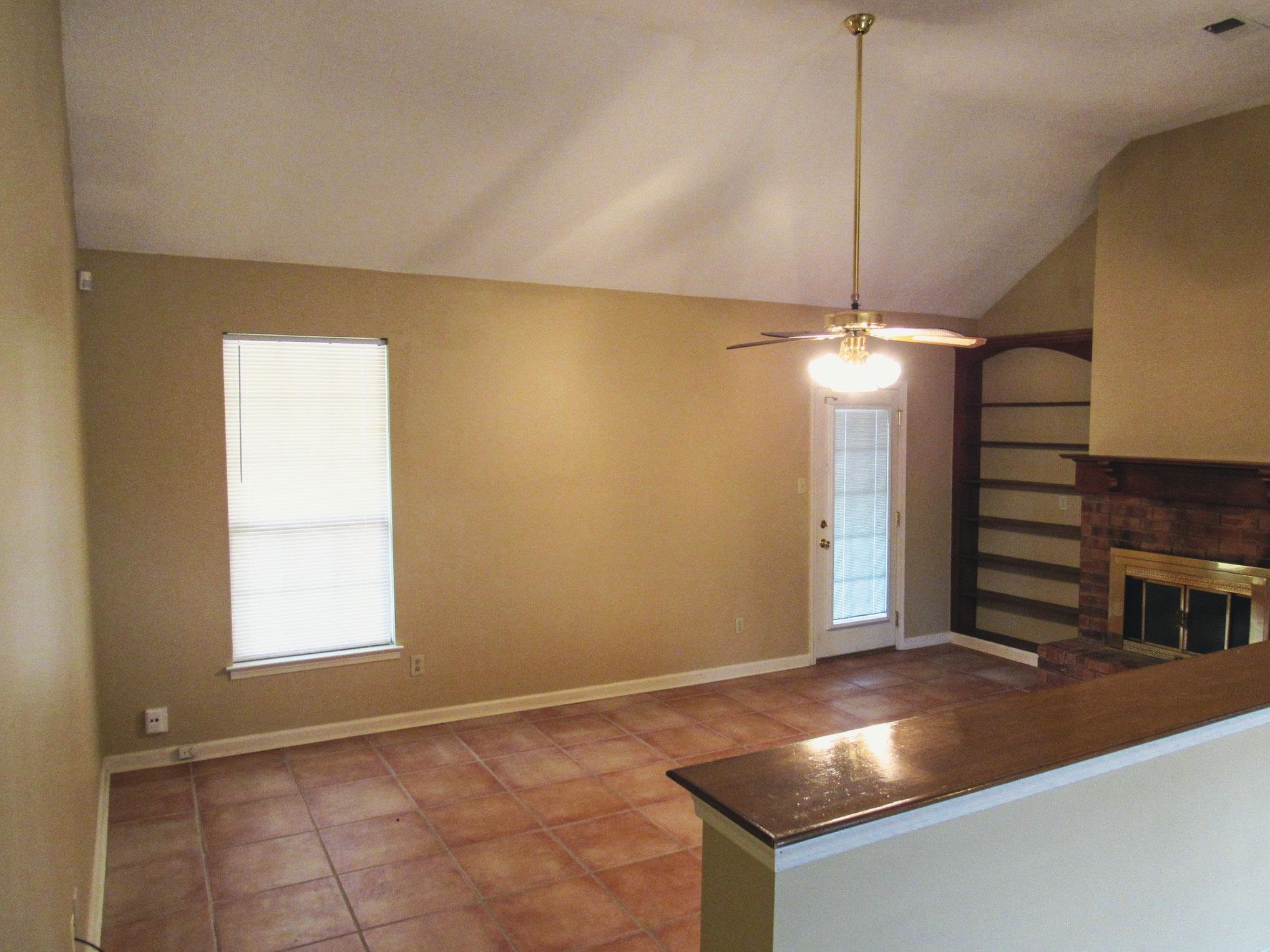 Empty living room with terracotta tile, a brick fireplace, built-in shelves, and a ceiling fan.