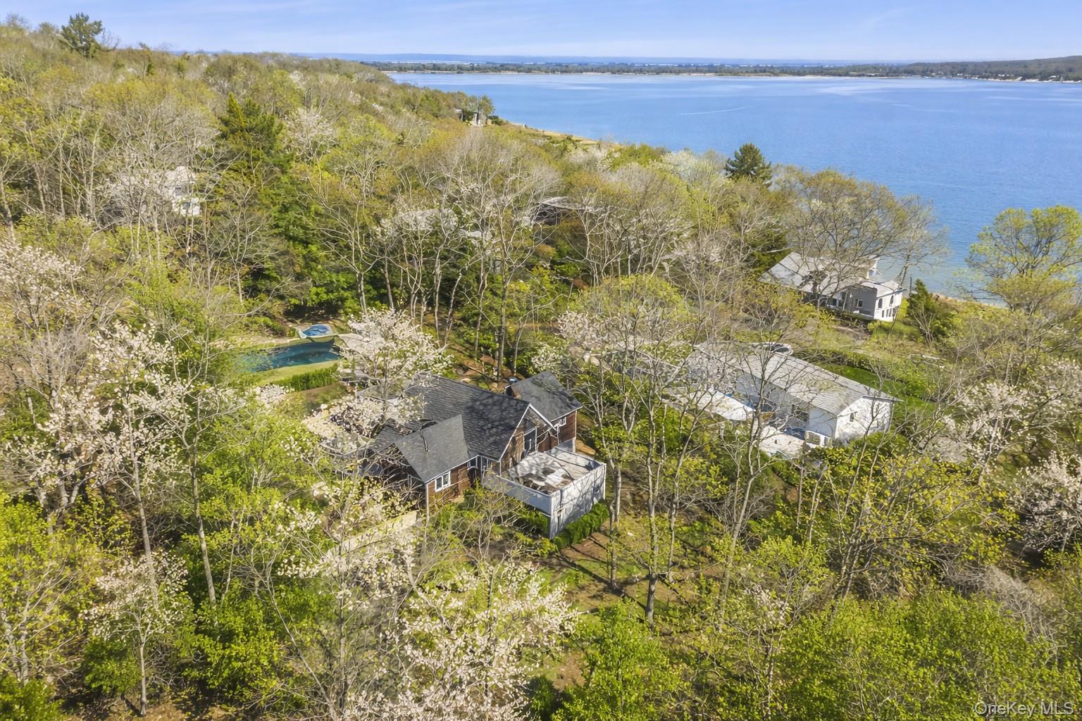 Aerial view of a wooded property with a swimming pool and bay beyond.