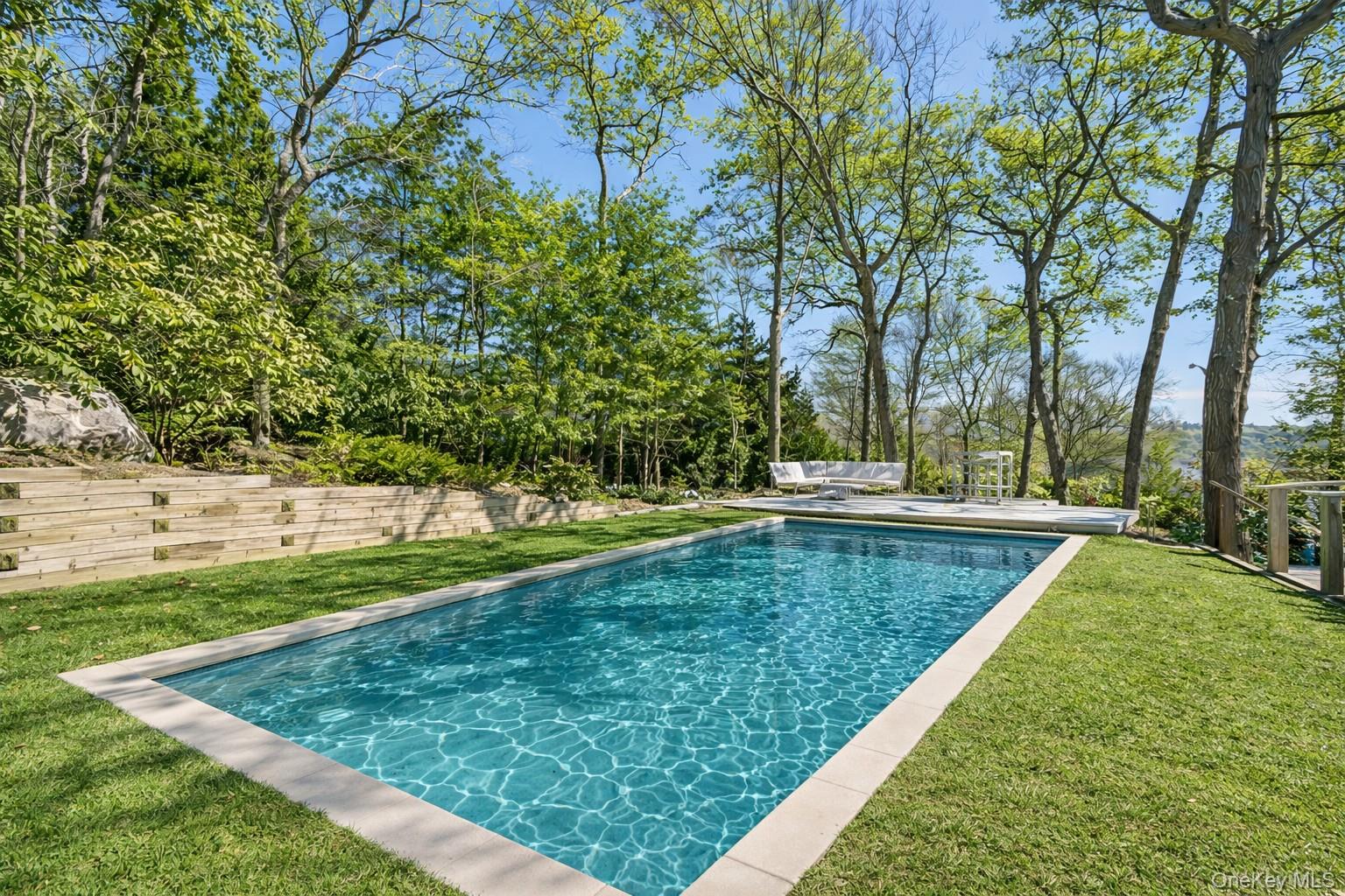 Rectangular swimming pool in a wooded yard, bordered by lawn and a low timber retaining wall.