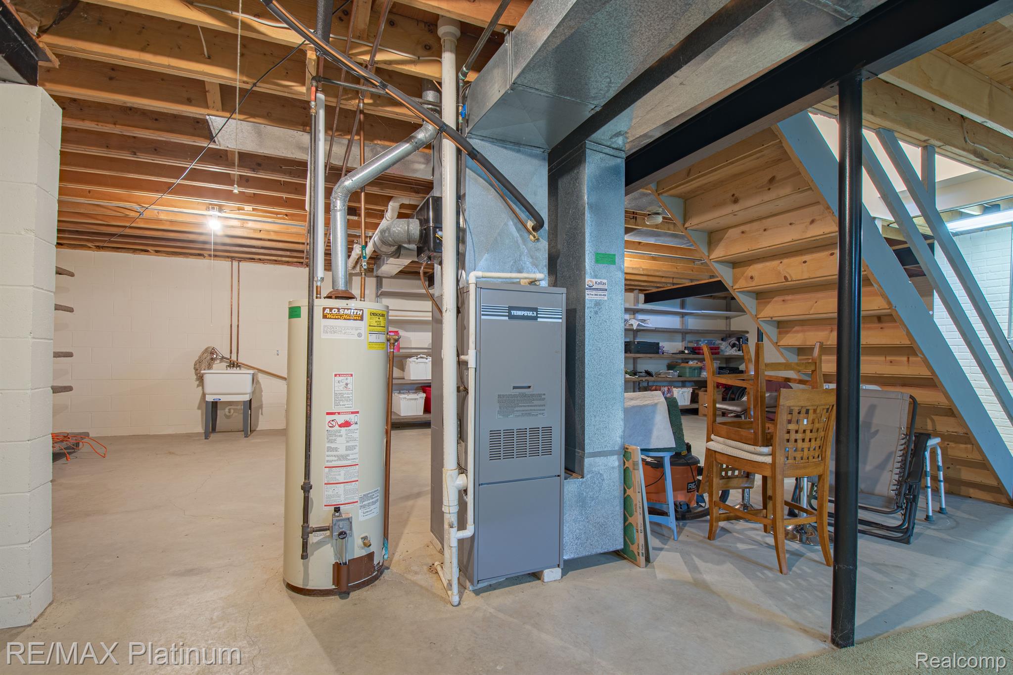 Basement utility area with a water heater and furnace beneath exposed joists, beside wooden stairs.