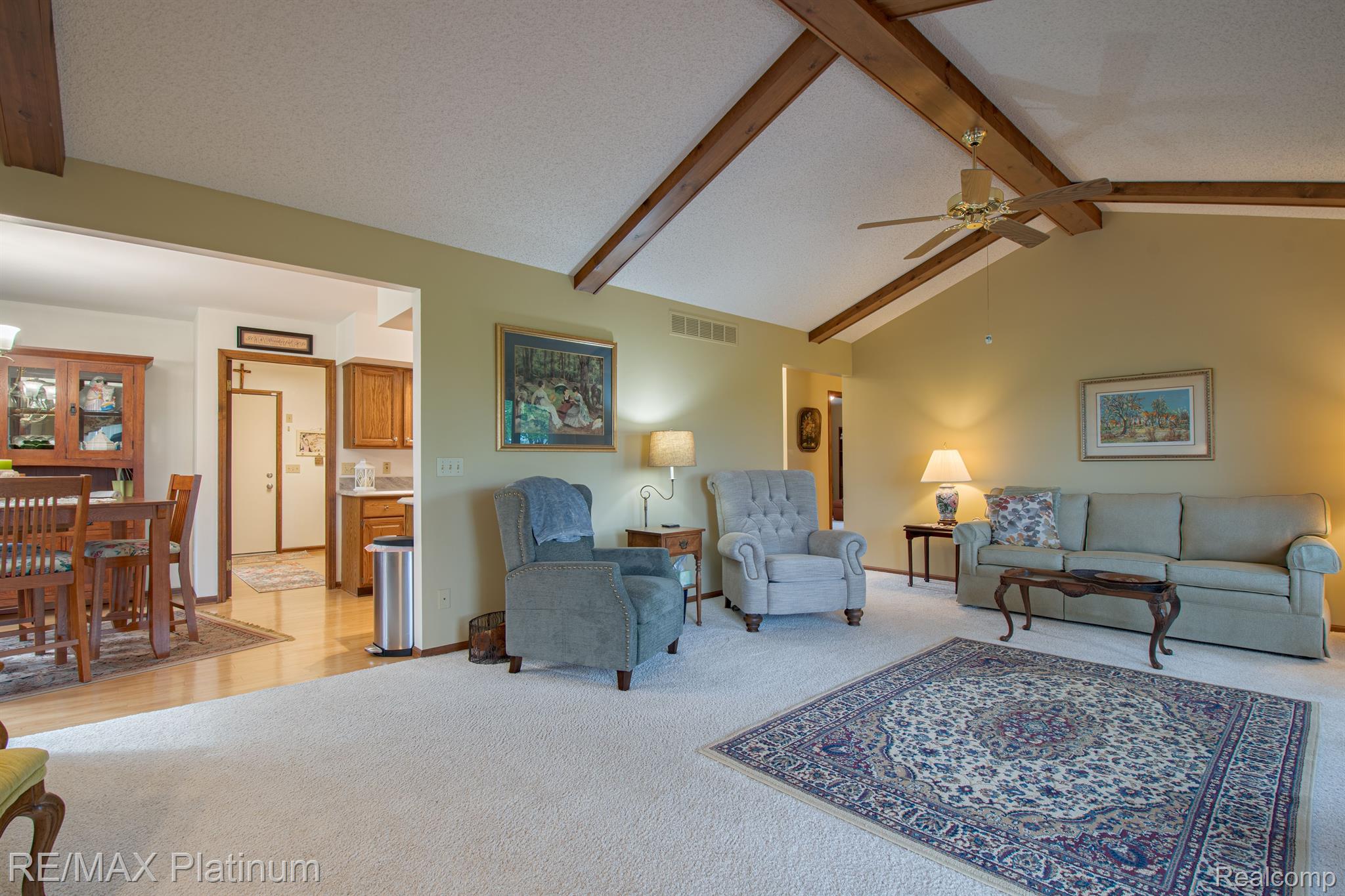 Living room with vaulted wood-beam ceiling, gray seating, patterned rug, and adjoining dining area.