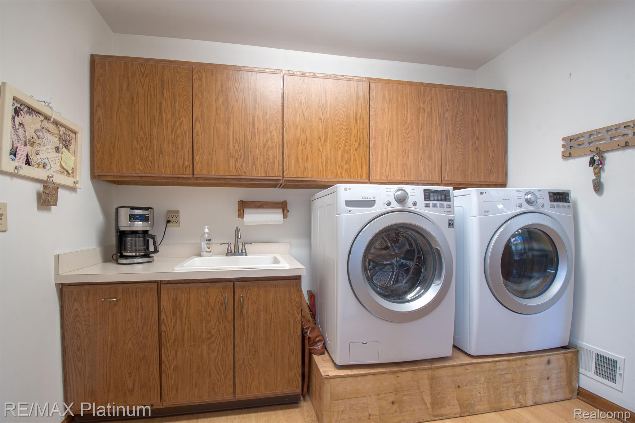 Laundry room with front-loading washer and dryer on a raised platform beside wood cabinets and a utility sink.