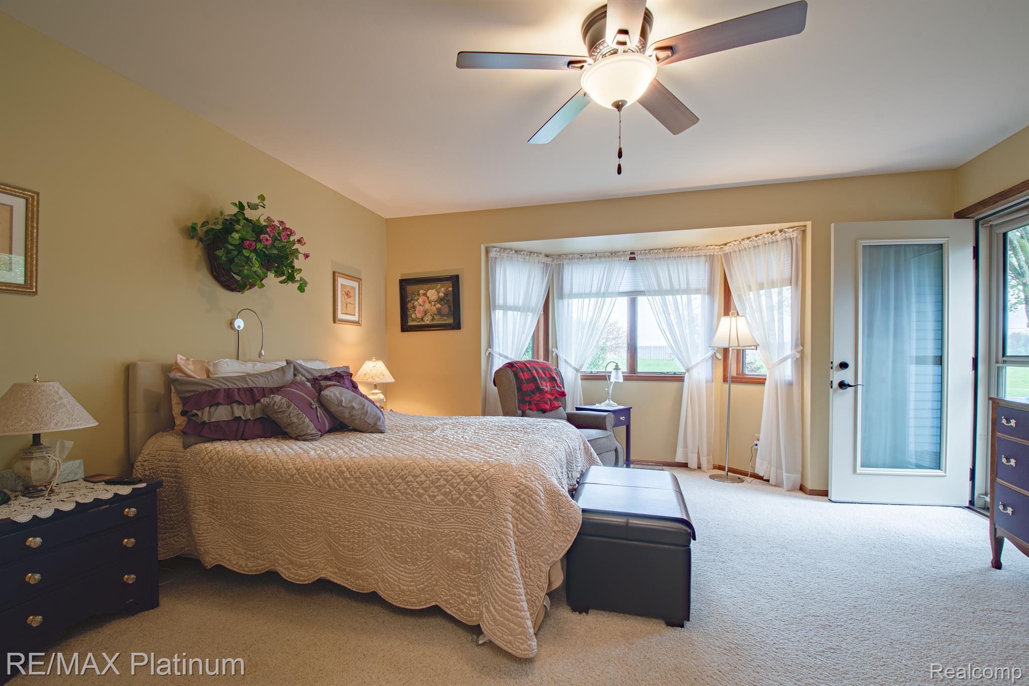 Furnished bedroom with a large quilted bed, ceiling fan, and curtained windows beside an open glass-panel door.
