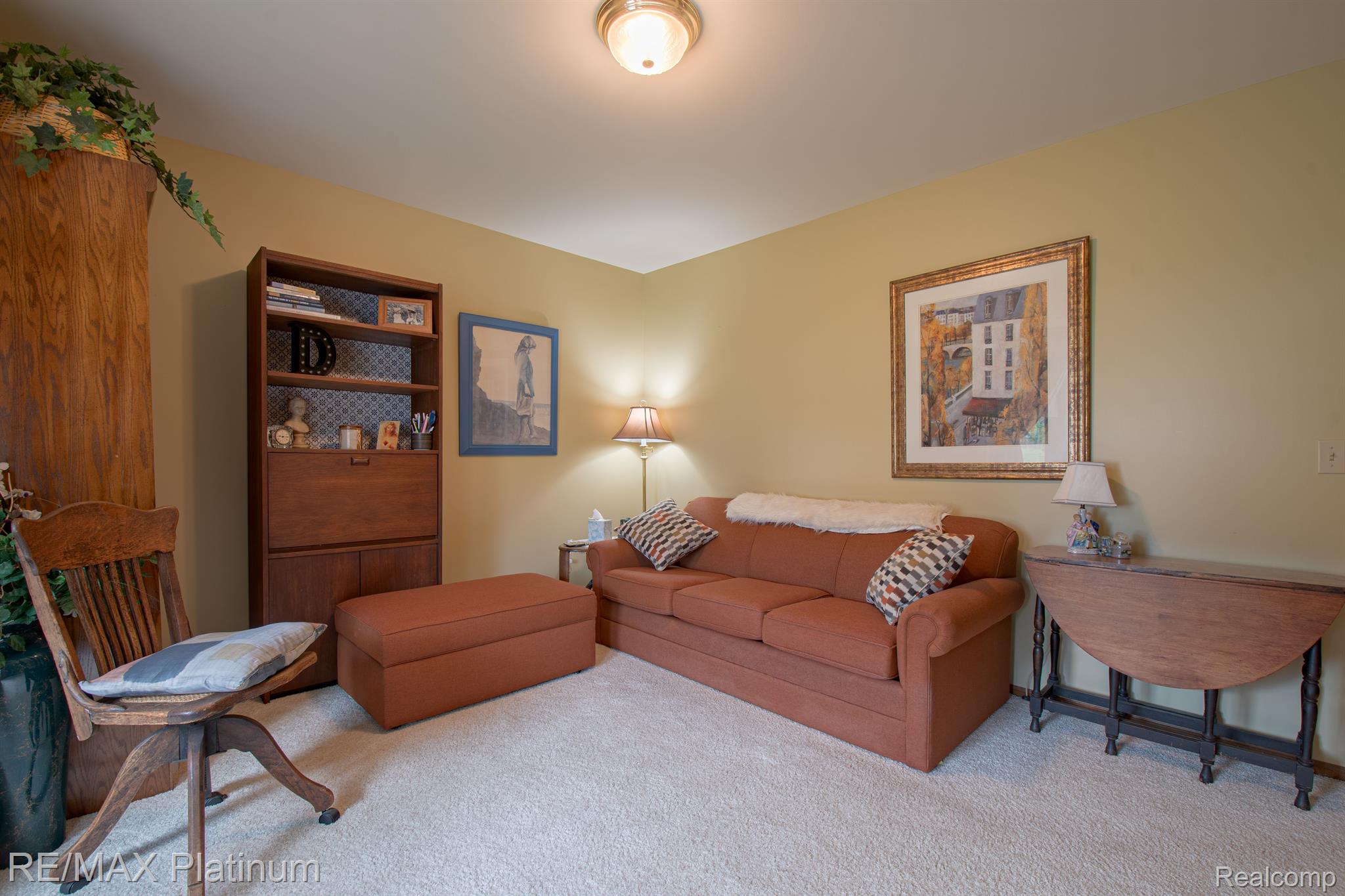 Furnished living room with rust-colored sofa, ottoman, wooden bookcase, and framed artwork.