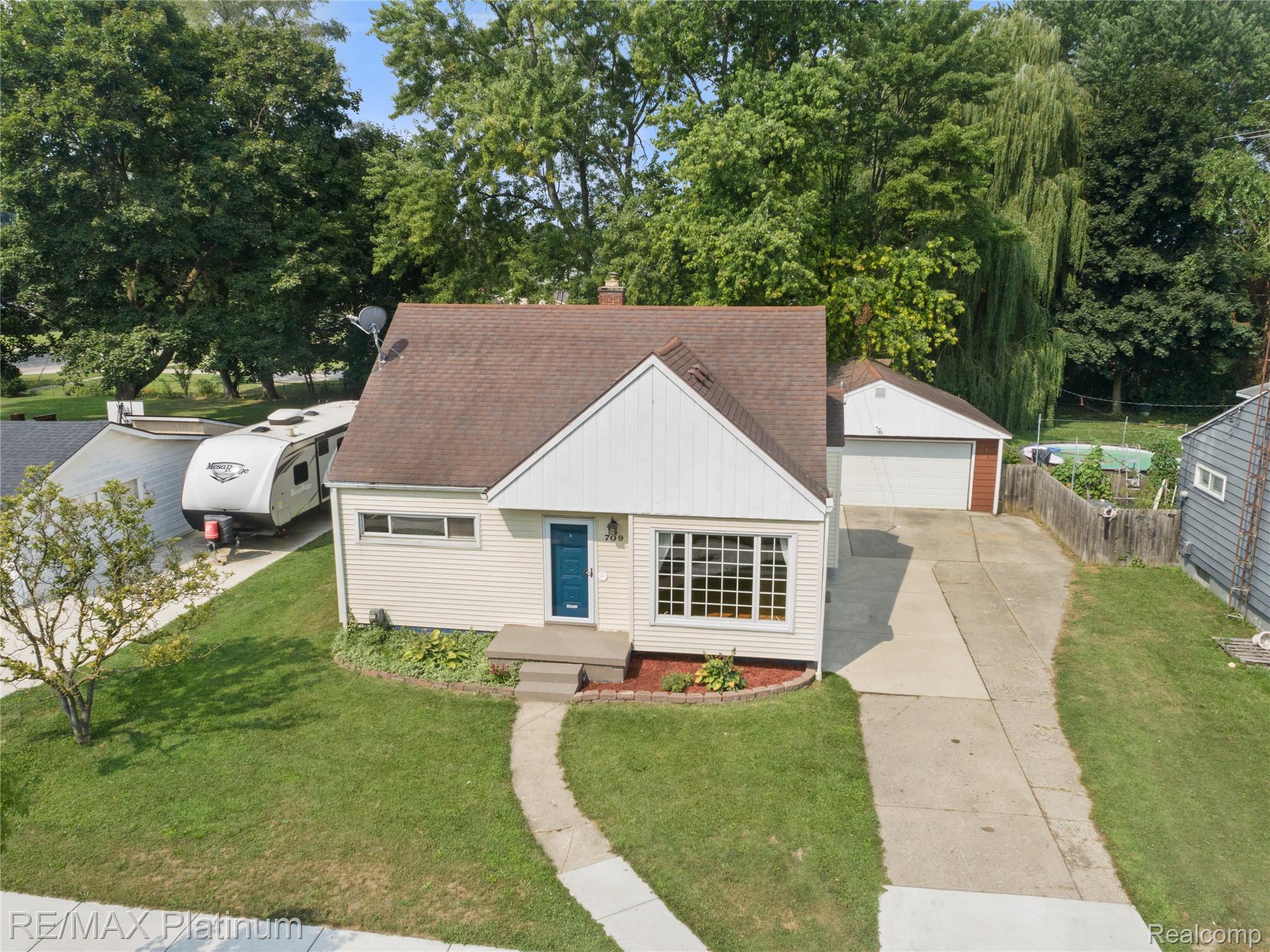 Aerial view of a small home with a blue front door, broad driveway, detached garage, and mature trees.