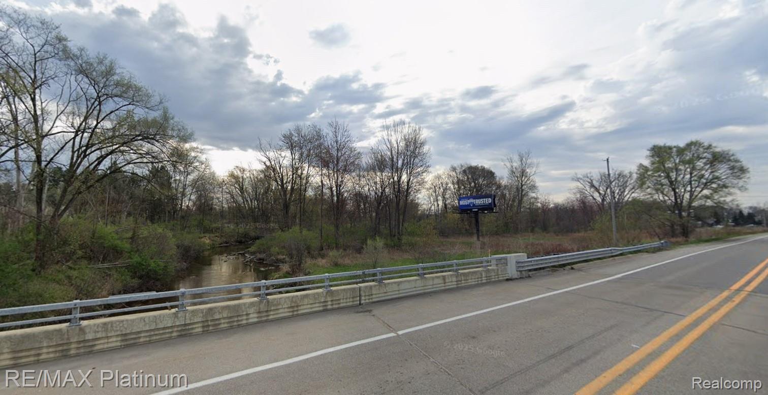 Wooded roadside land with a creek beneath a bridge beside a two-lane road.
