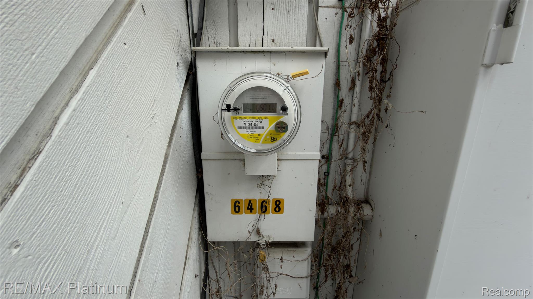 Exterior electric meter on white siding, with address number 6468 below it and dried vines alongside.