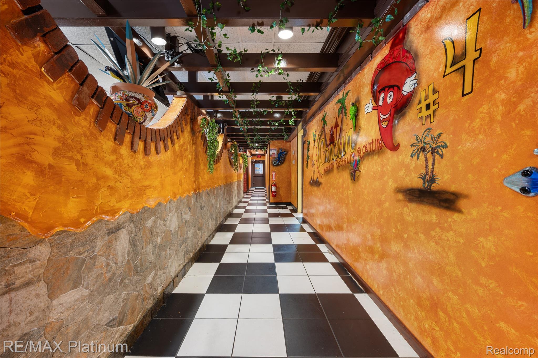 Commercial corridor with orange Mexican-themed murals, a checkerboard floor, stone walls, and trailing ceiling greenery.