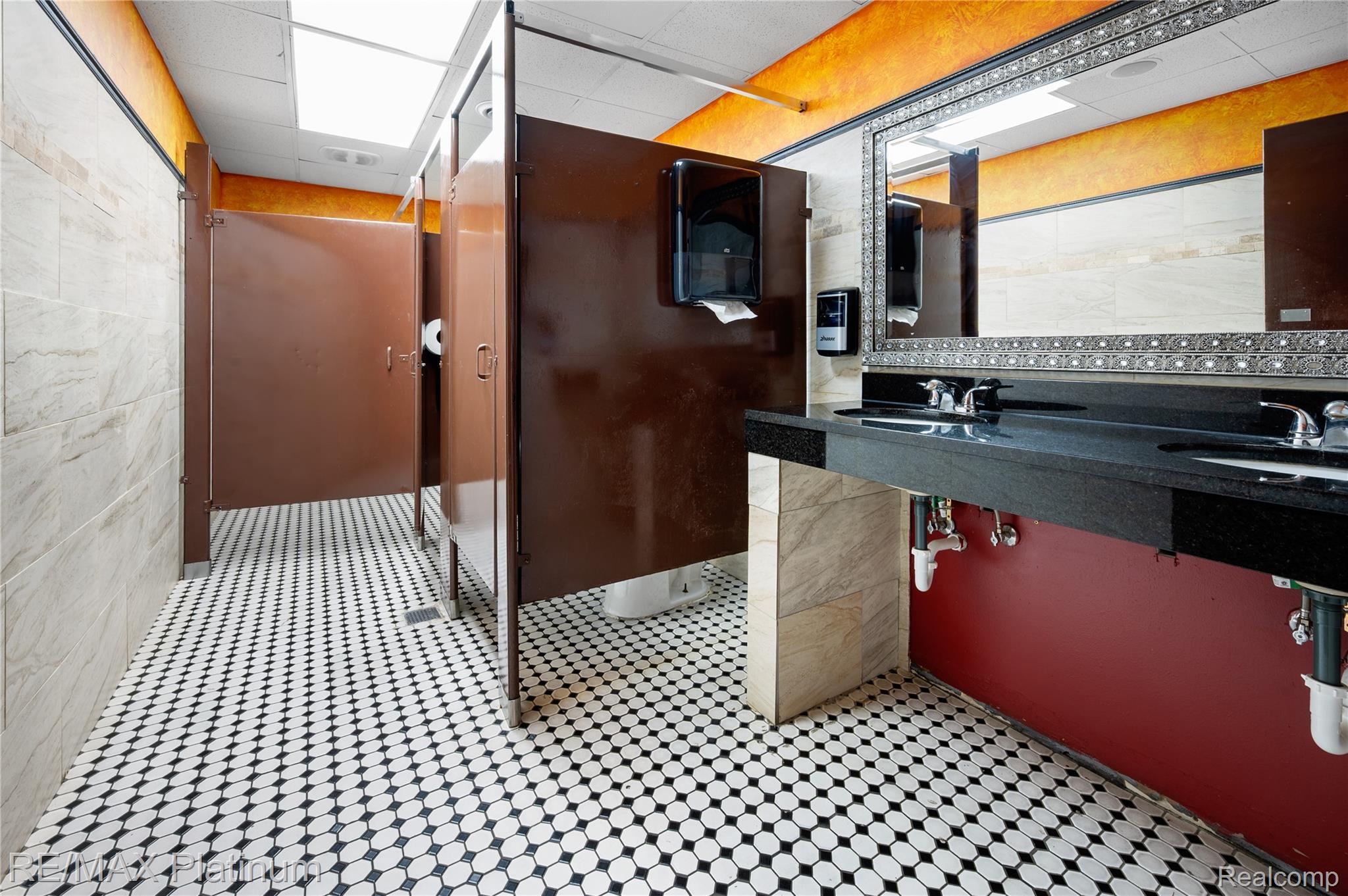 Commercial restroom with brown stalls, patterned tile floor, and a long double-sink vanity.