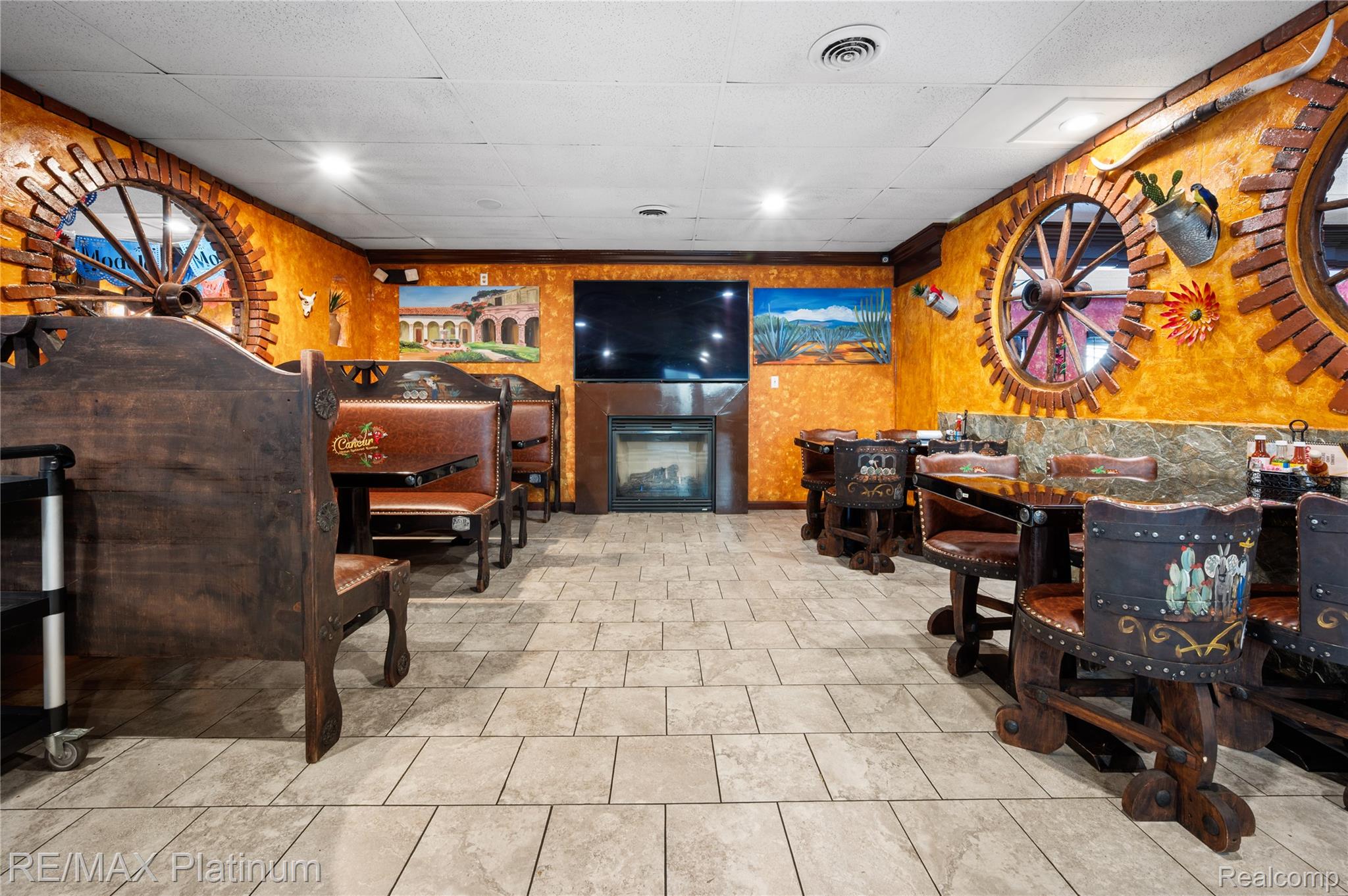 Restaurant dining area with orange walls, wagon-wheel windows, wooden booths, and a fireplace beneath a television.