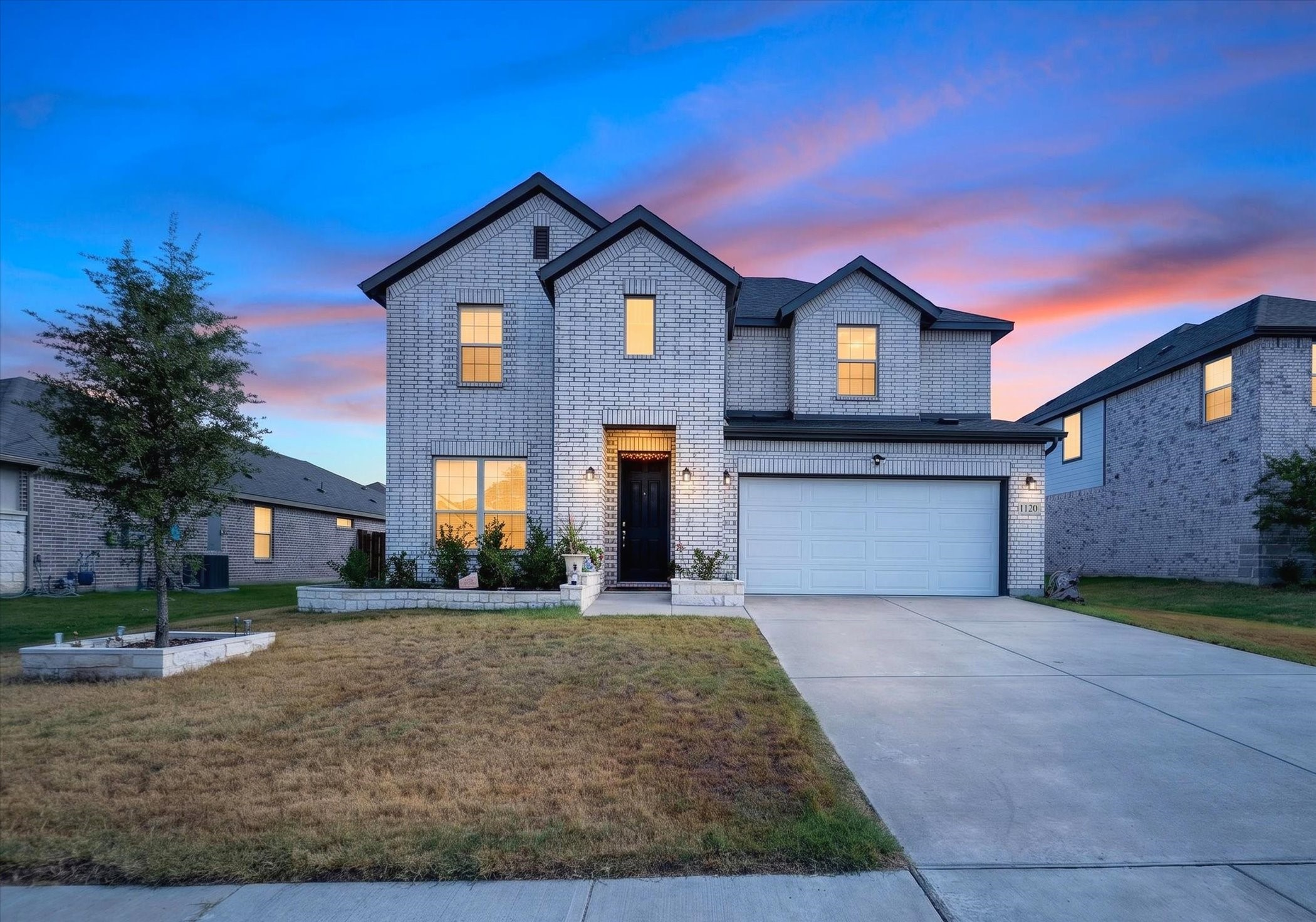 Two-story light-brick home with a two-car garage and illuminated windows at dusk.