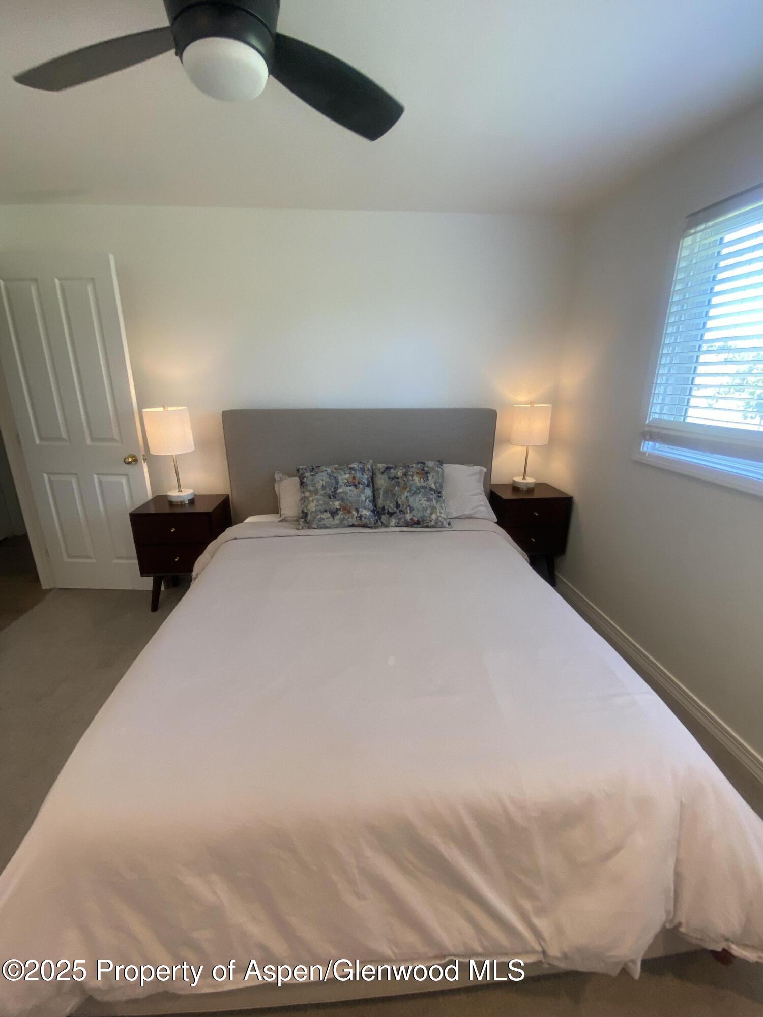 Bedroom with a neatly made bed, matching nightstands, lit lamps, and a ceiling fan.