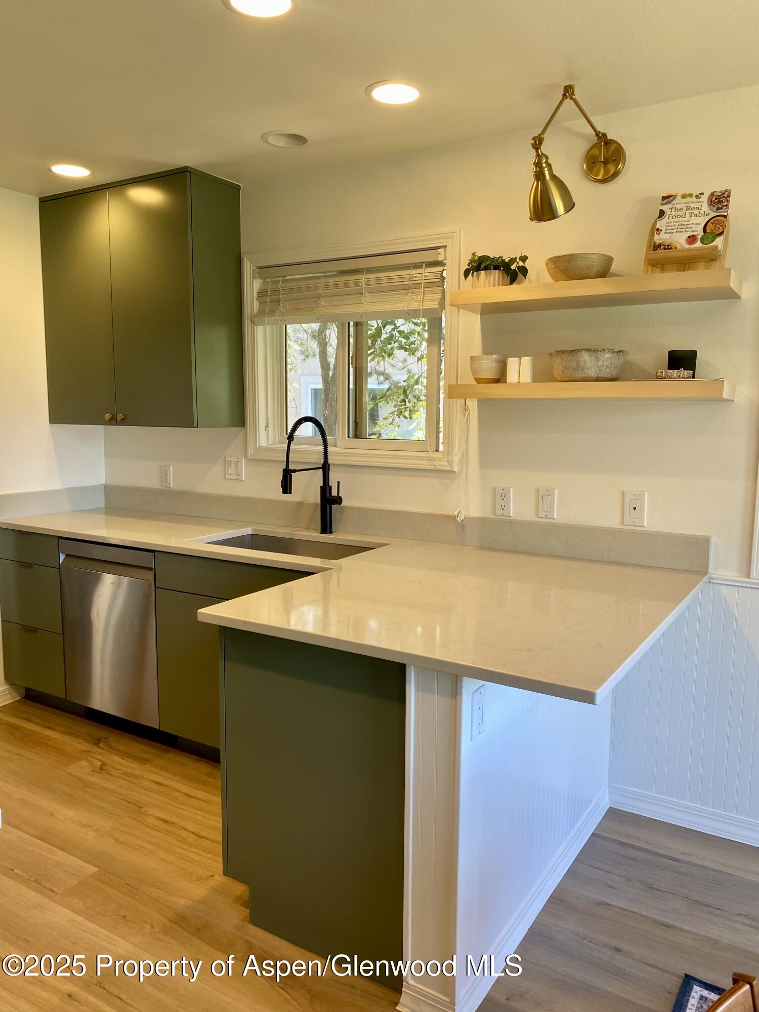 Kitchen with olive-green cabinets, white quartz peninsula, black faucet, and open shelves beside a window.