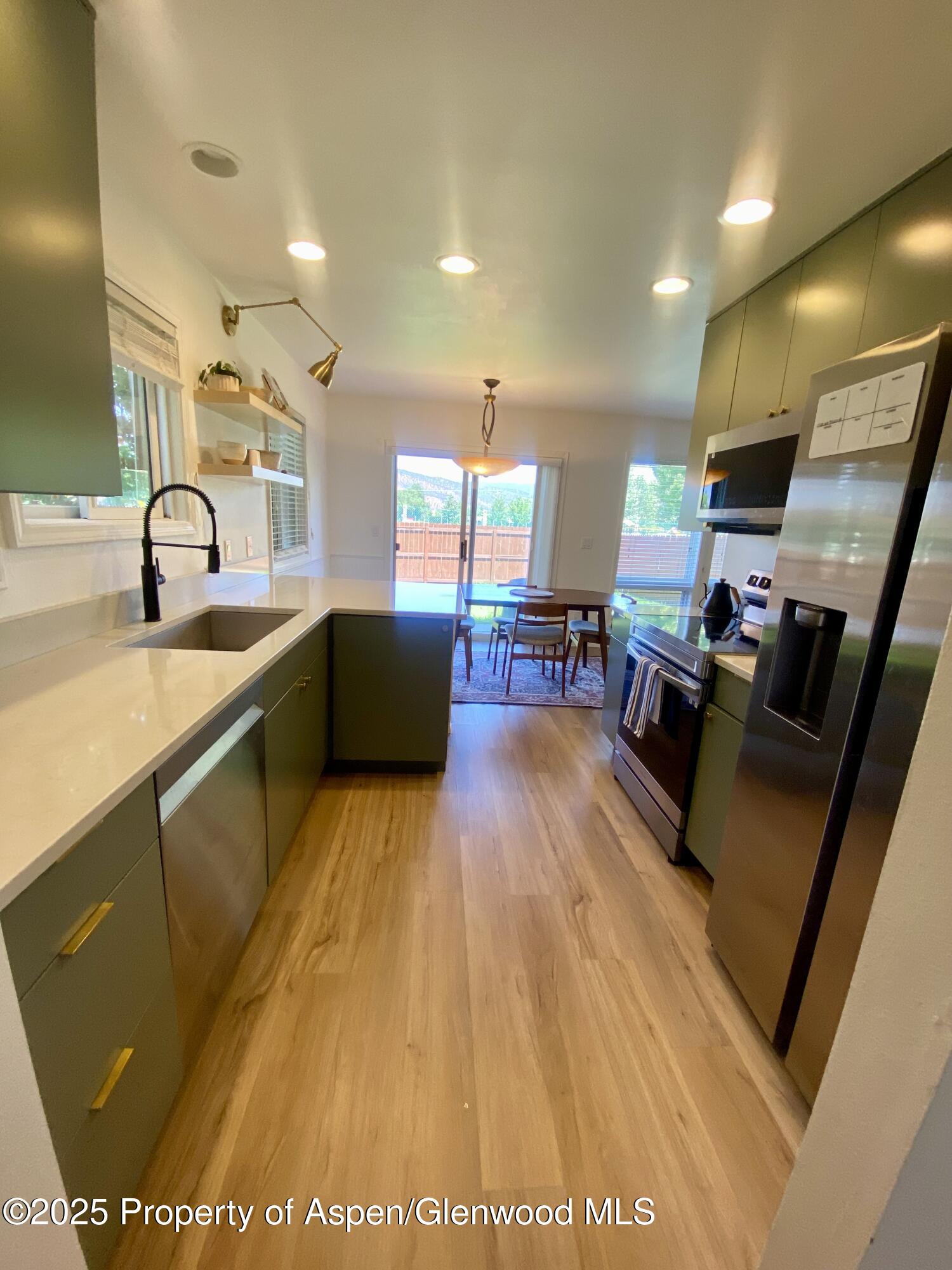 Modern kitchen with olive-green cabinets, white counters, stainless steel appliances, and a dining area by sliding glass doors.