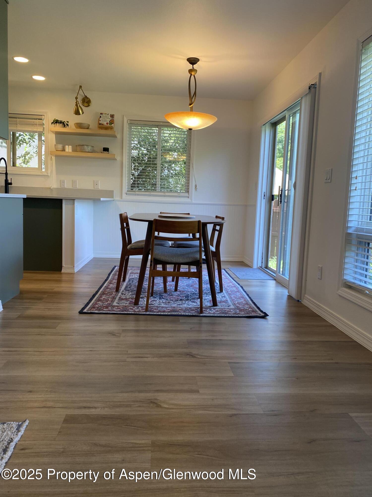 Dining area with a round wooden table, patterned rug, open kitchen shelves, and sliding glass doors.