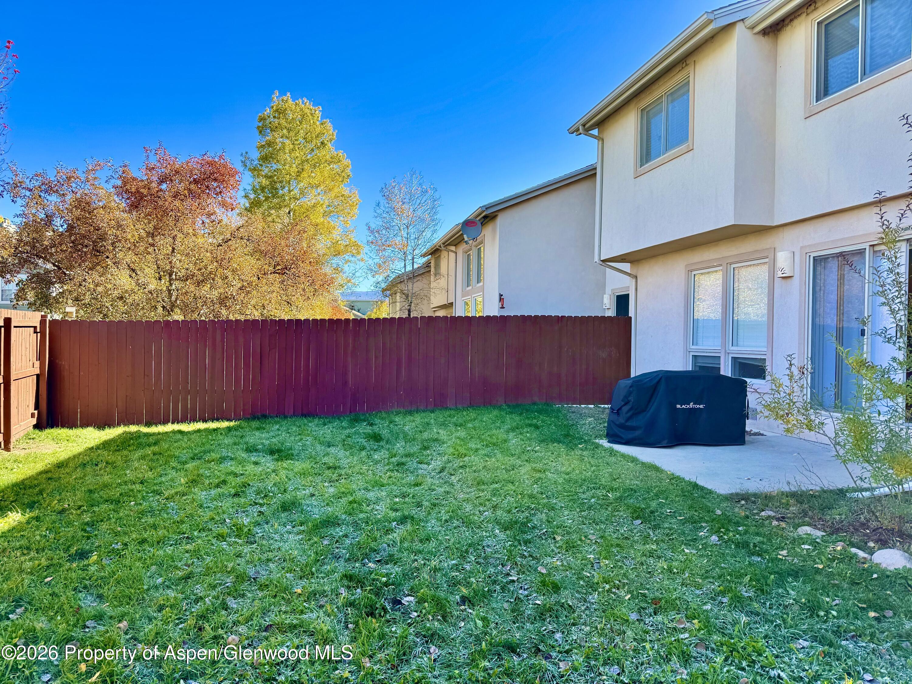 Fenced backyard with a broad lawn, autumn trees, and a covered grill on the patio.