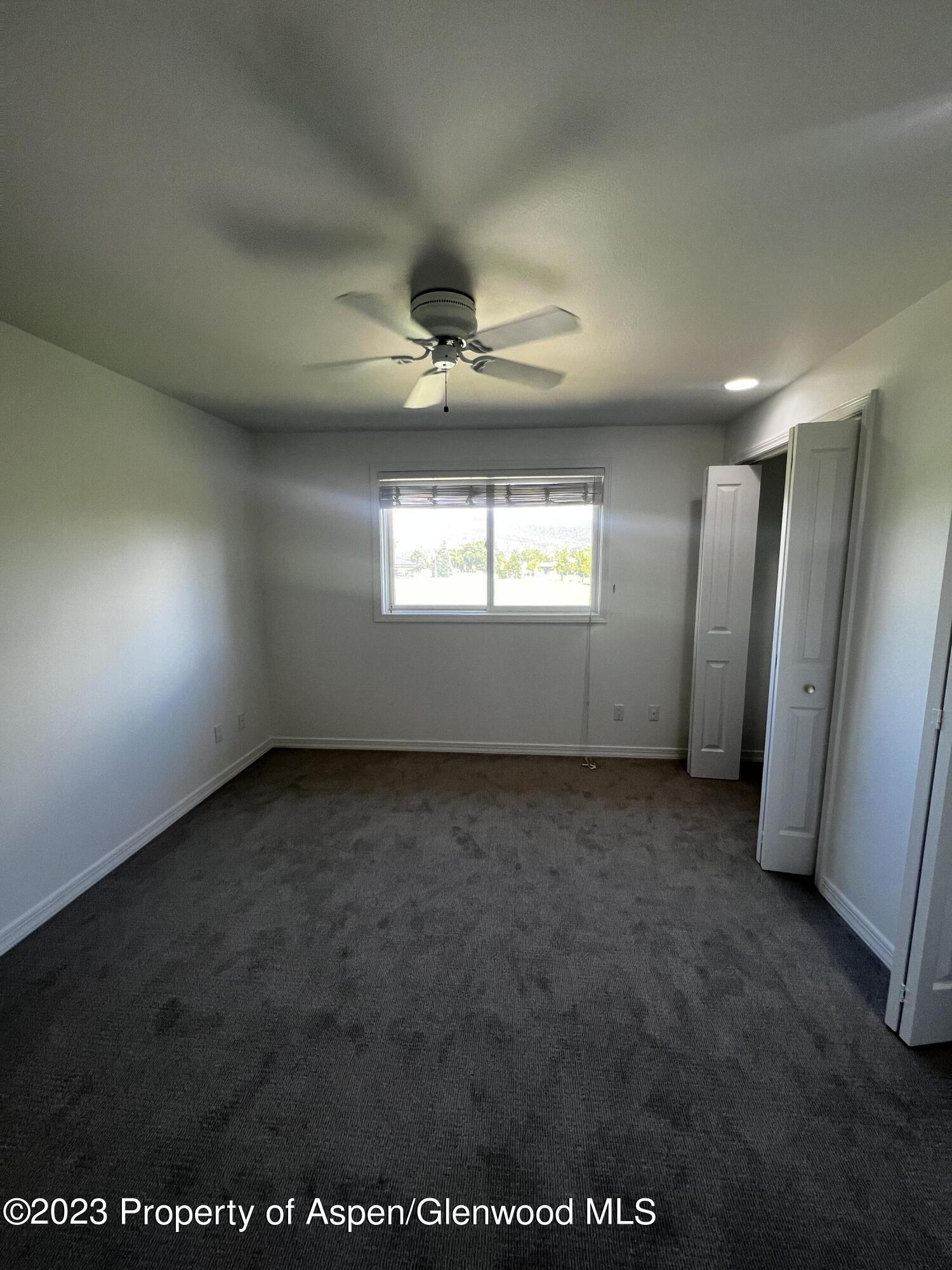 Empty bedroom with gray carpet, a ceiling fan, window with blinds, and open closet doors.