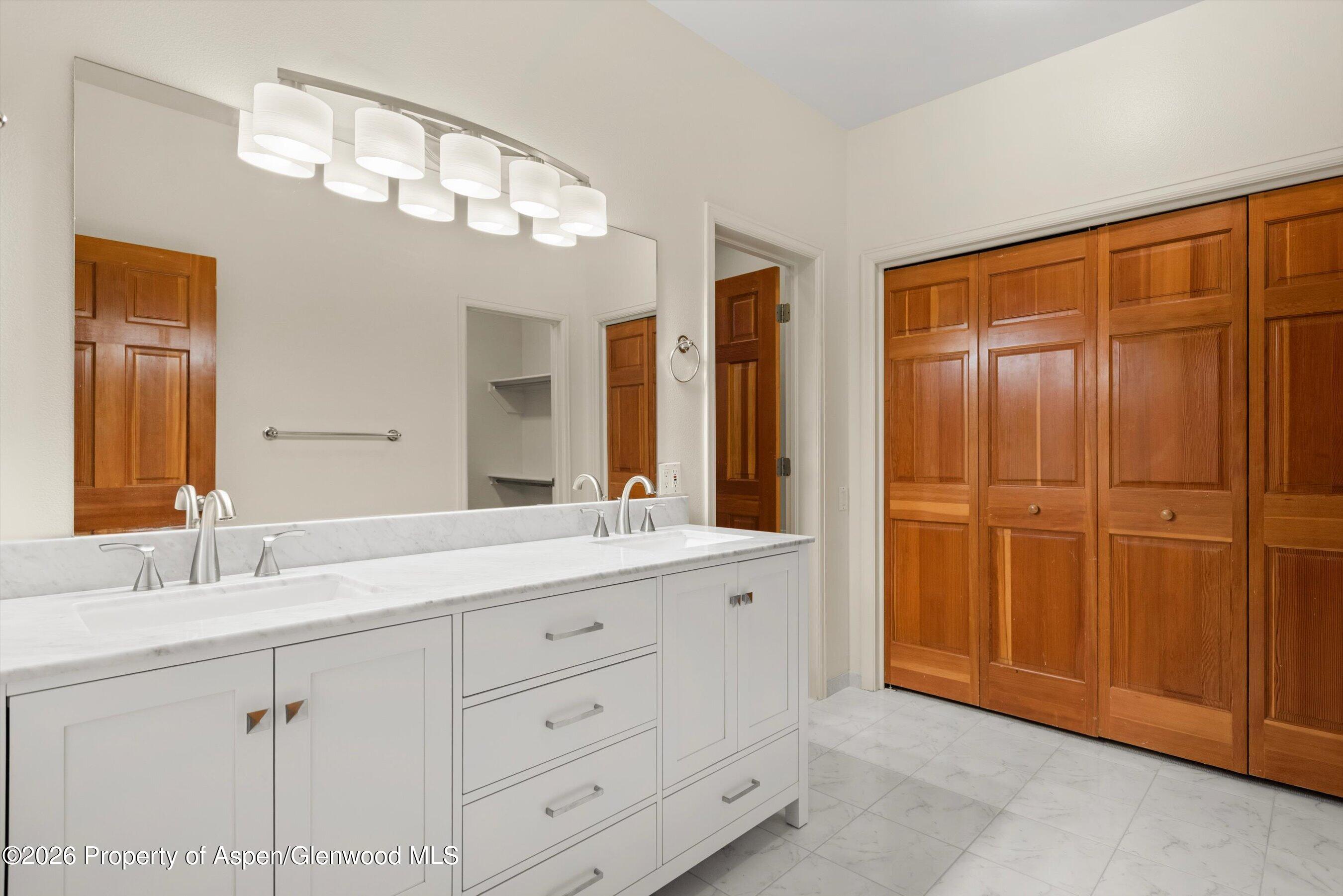 Bathroom with a white double vanity and marble surfaces beside warm wood closet doors.