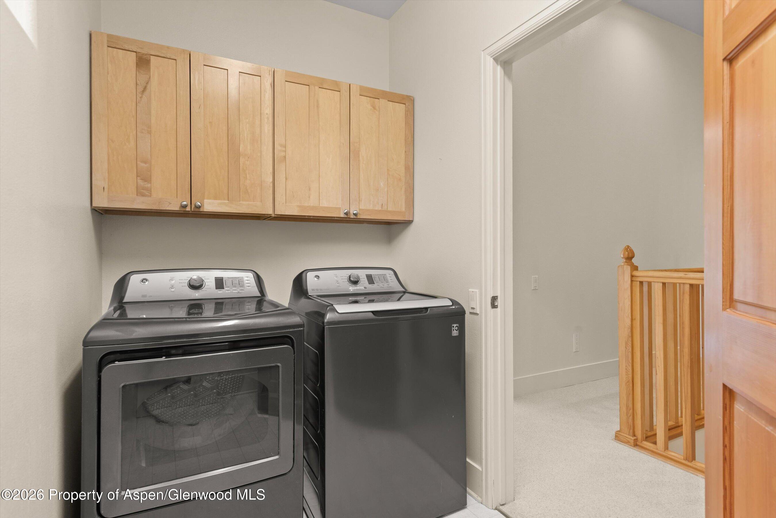 Laundry room with dark washer and dryer beneath light wood cabinets, beside a carpeted landing.