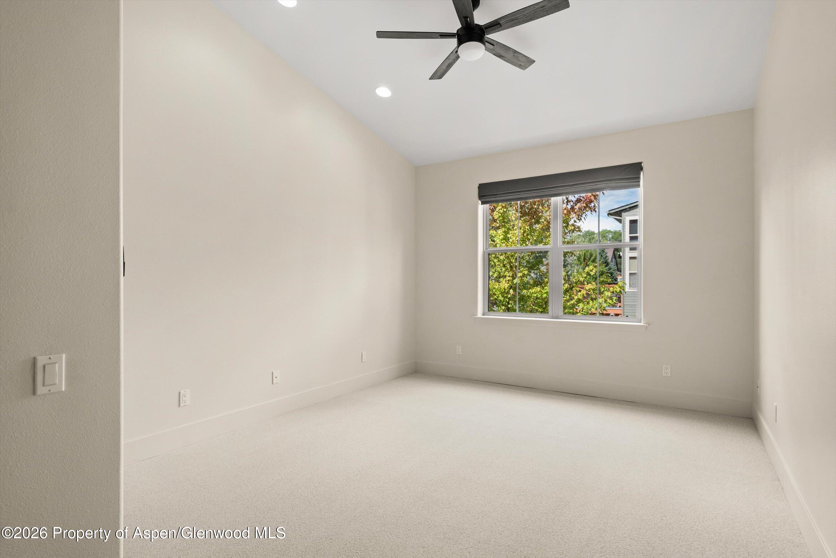 Empty carpeted bedroom with a black ceiling fan and a window overlooking leafy trees.