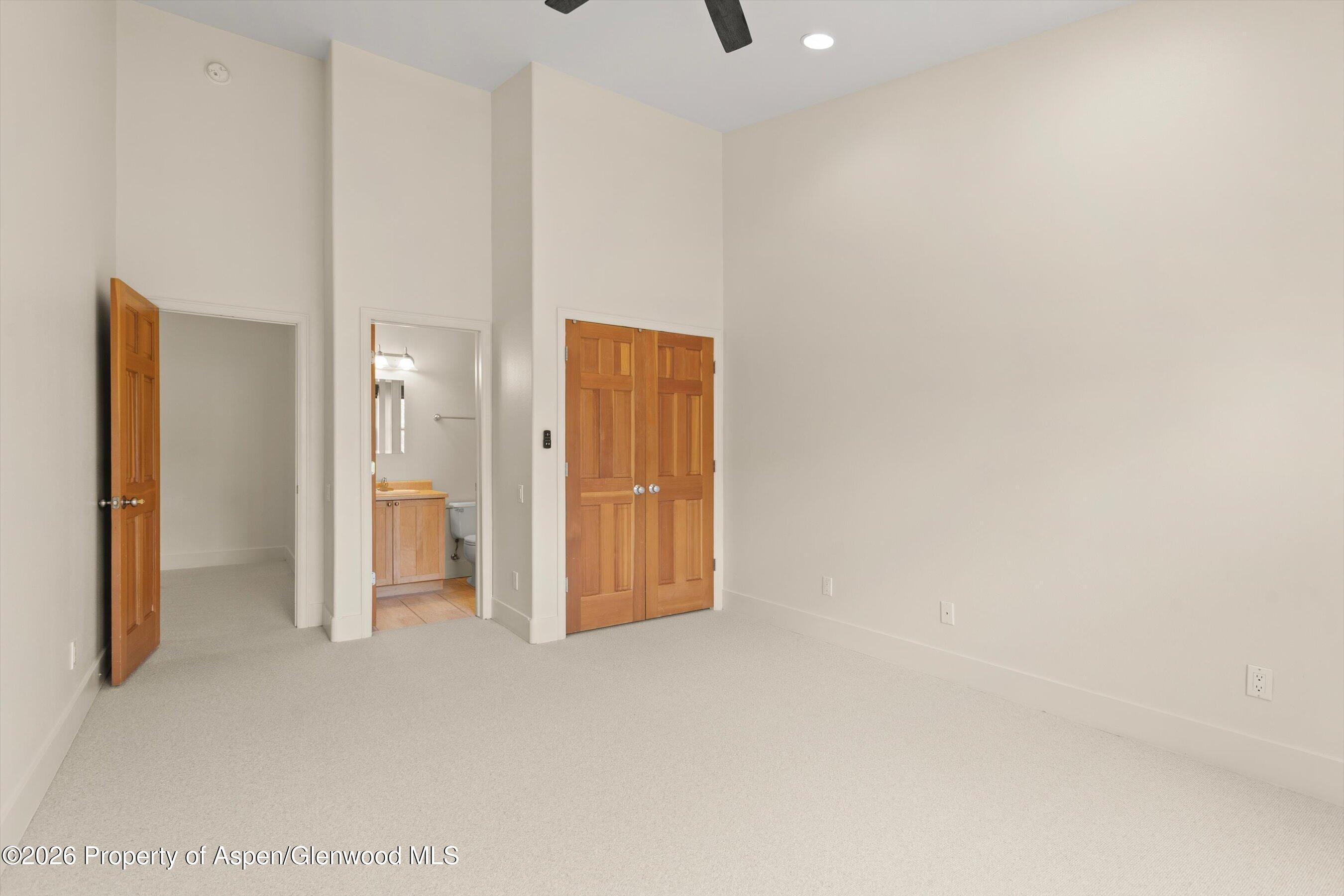 Empty carpeted bedroom with a high ceiling and wooden doors beside an open bathroom doorway.