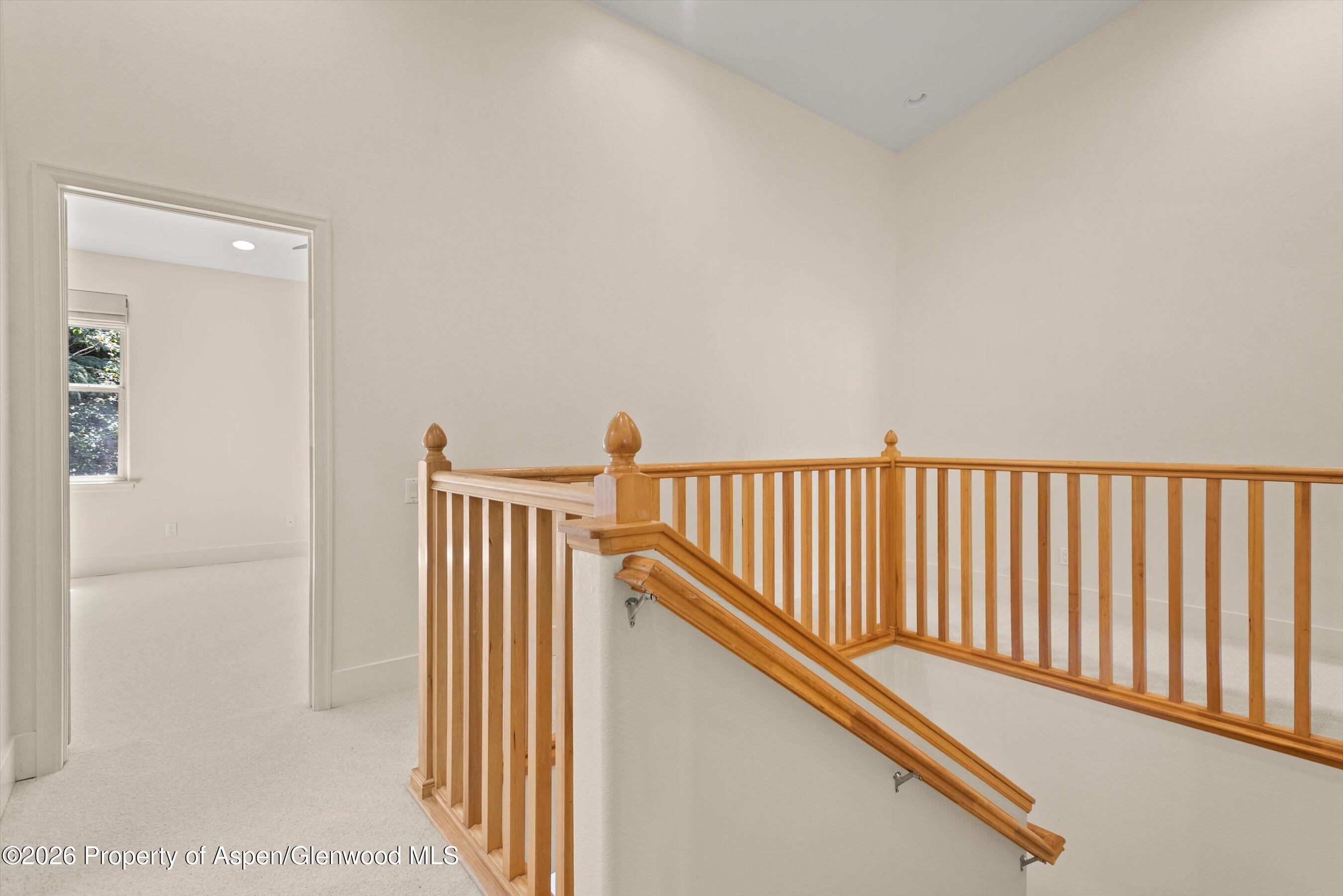 Upstairs landing with a wooden stair railing, carpeted floor, and doorway to a bright room.