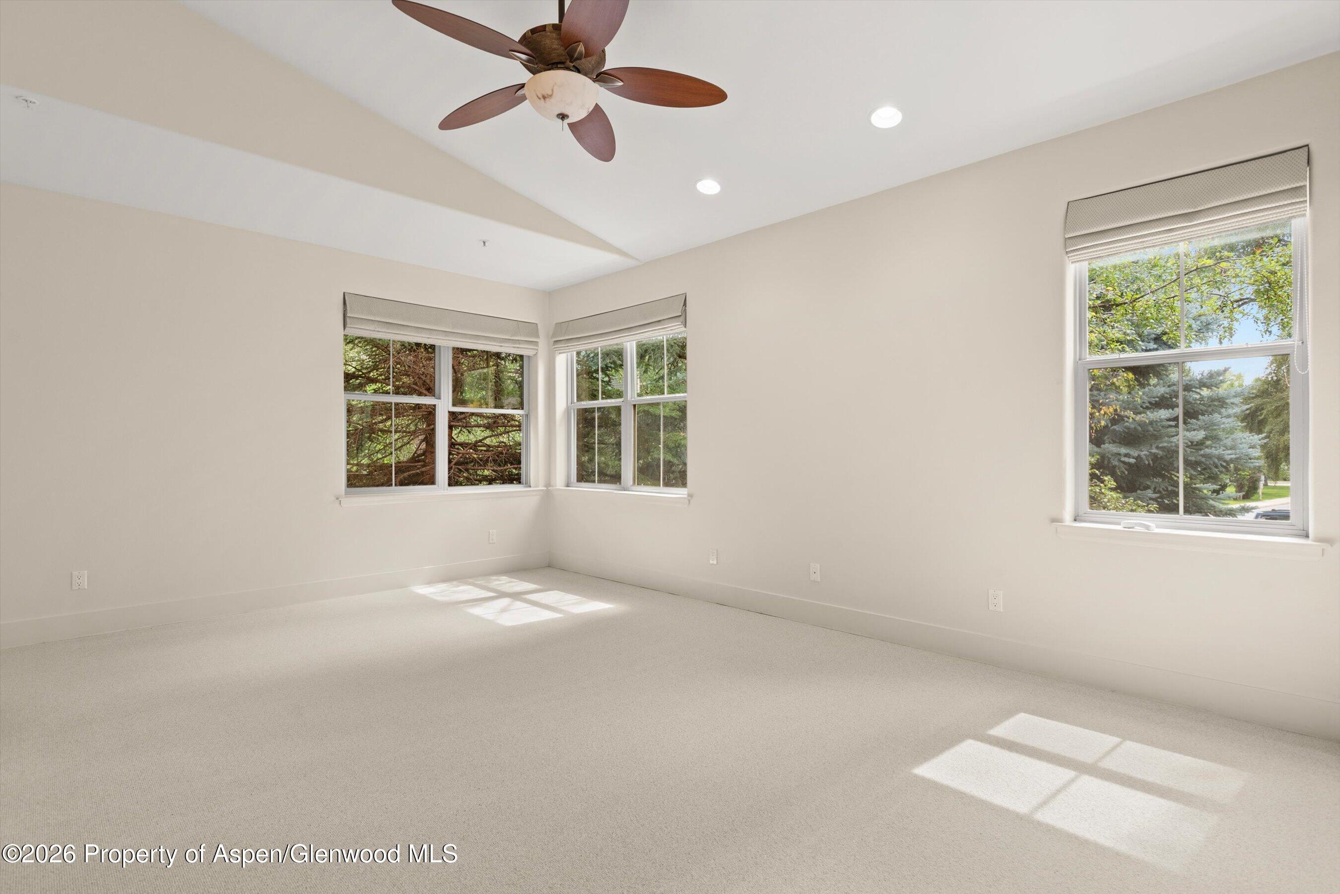 Empty carpeted bedroom with a ceiling fan and three windows overlooking trees.