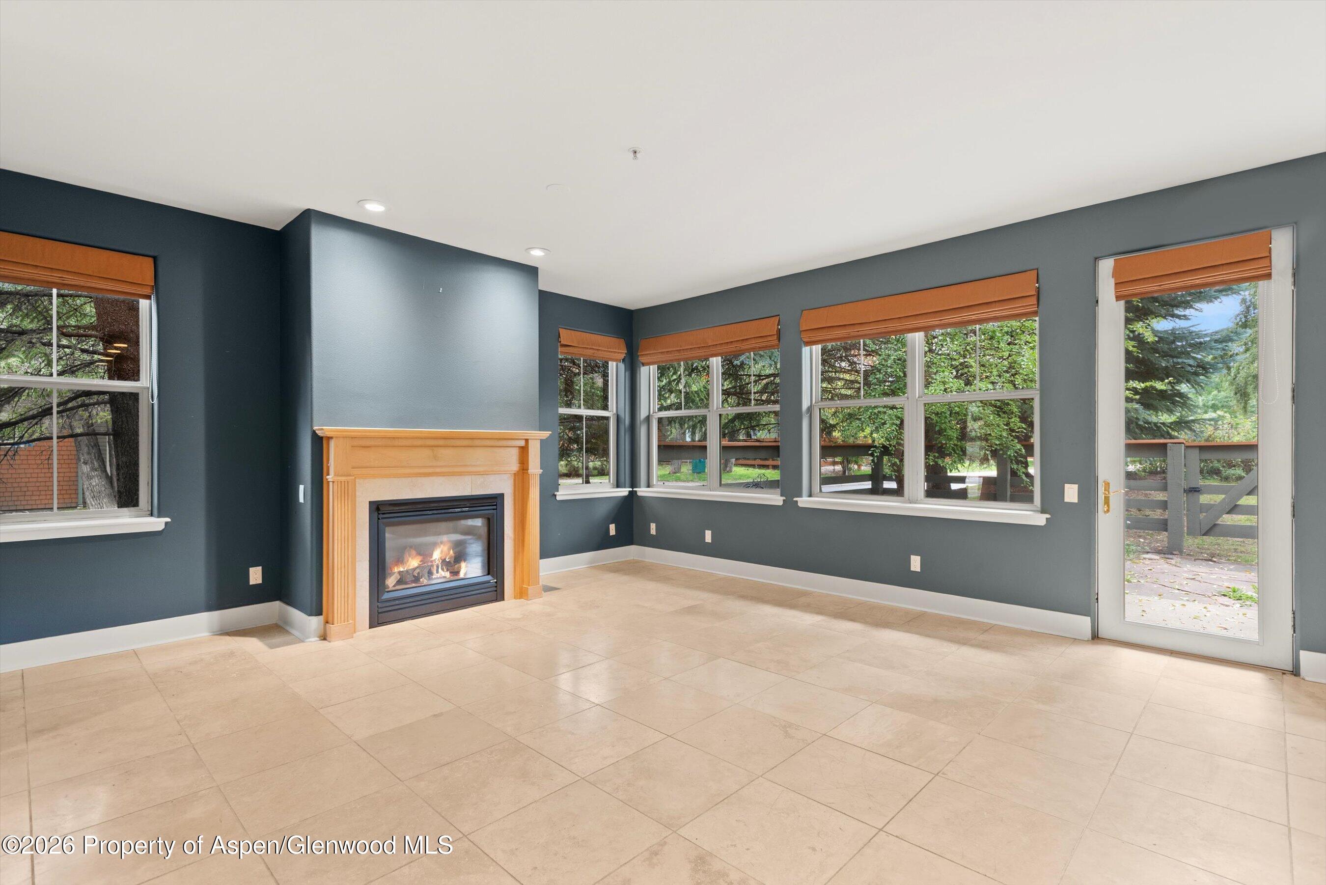 Vacant living room with a wood-trimmed fireplace, tiled floor, and windows overlooking trees.