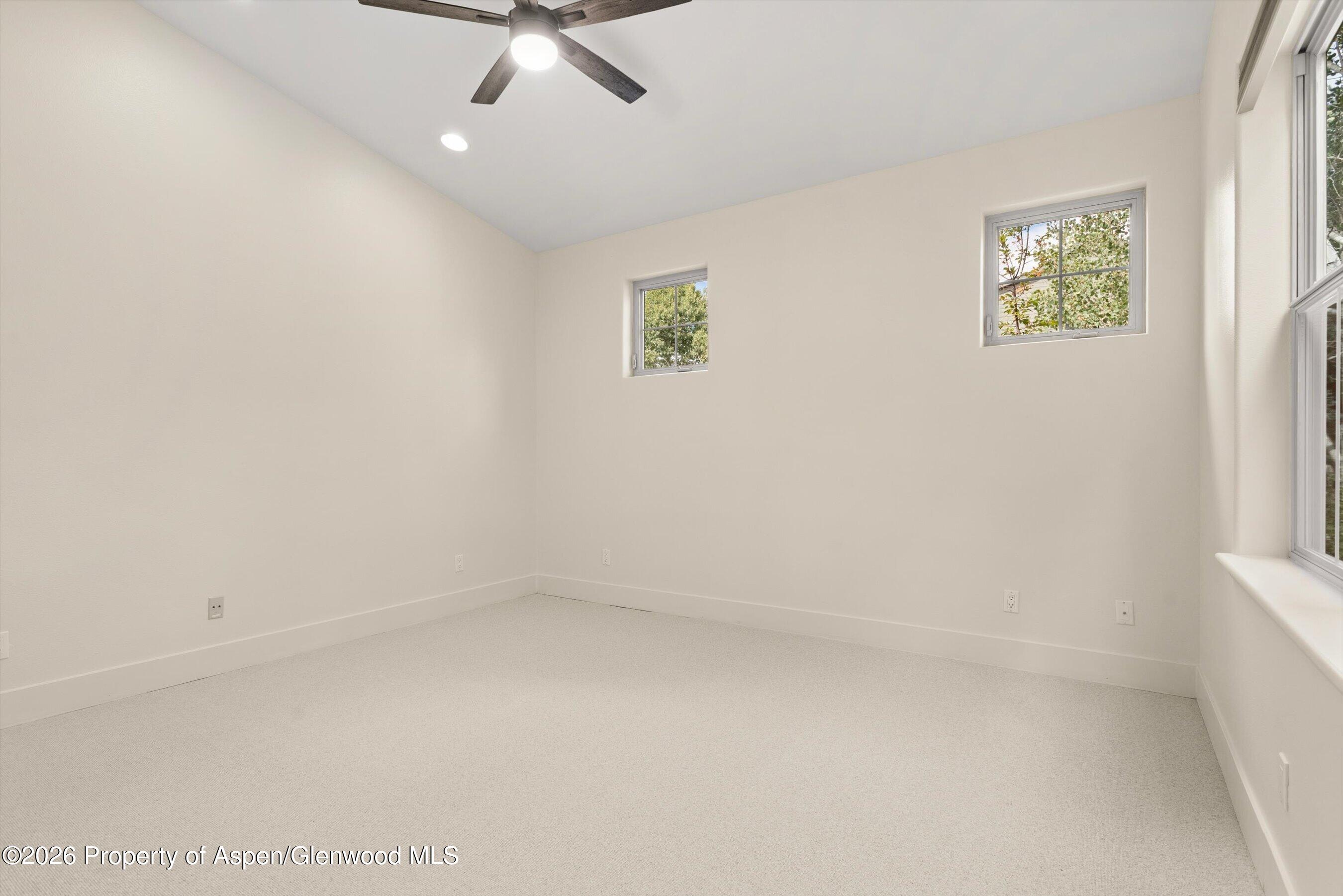 Empty carpeted bedroom with a ceiling fan and windows overlooking trees.