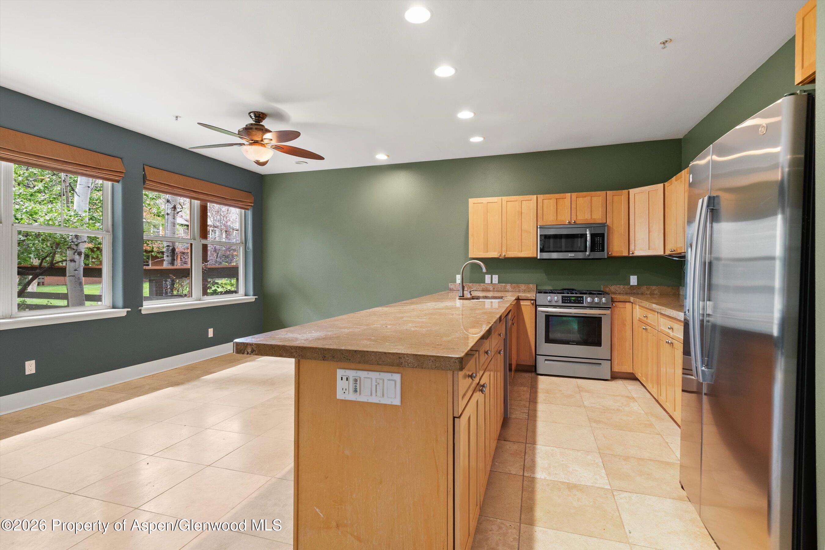 Kitchen with a large stone-topped island, wood cabinetry, stainless steel appliances, and windows overlooking trees.