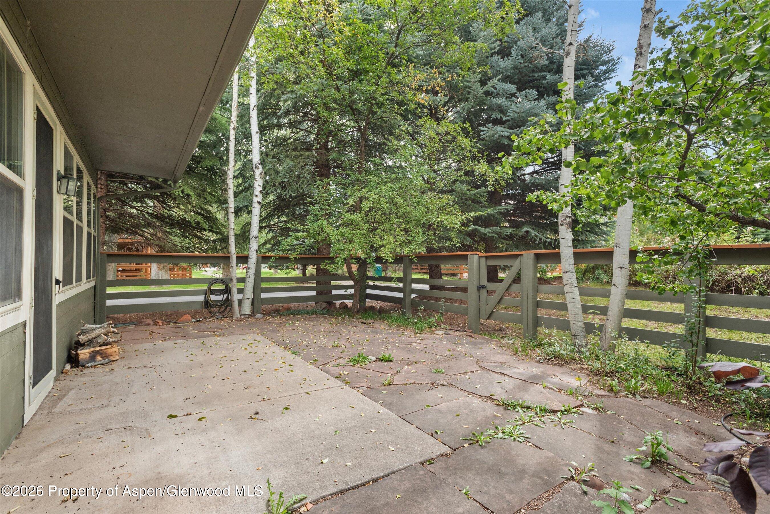 Concrete patio beside the home, shaded by a roof overhang and bordered by a wooden fence and trees.