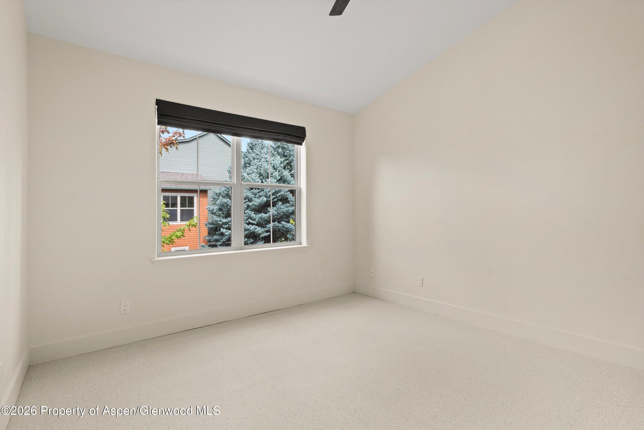 Empty carpeted room with a black-shaded window overlooking trees and a neighboring building.