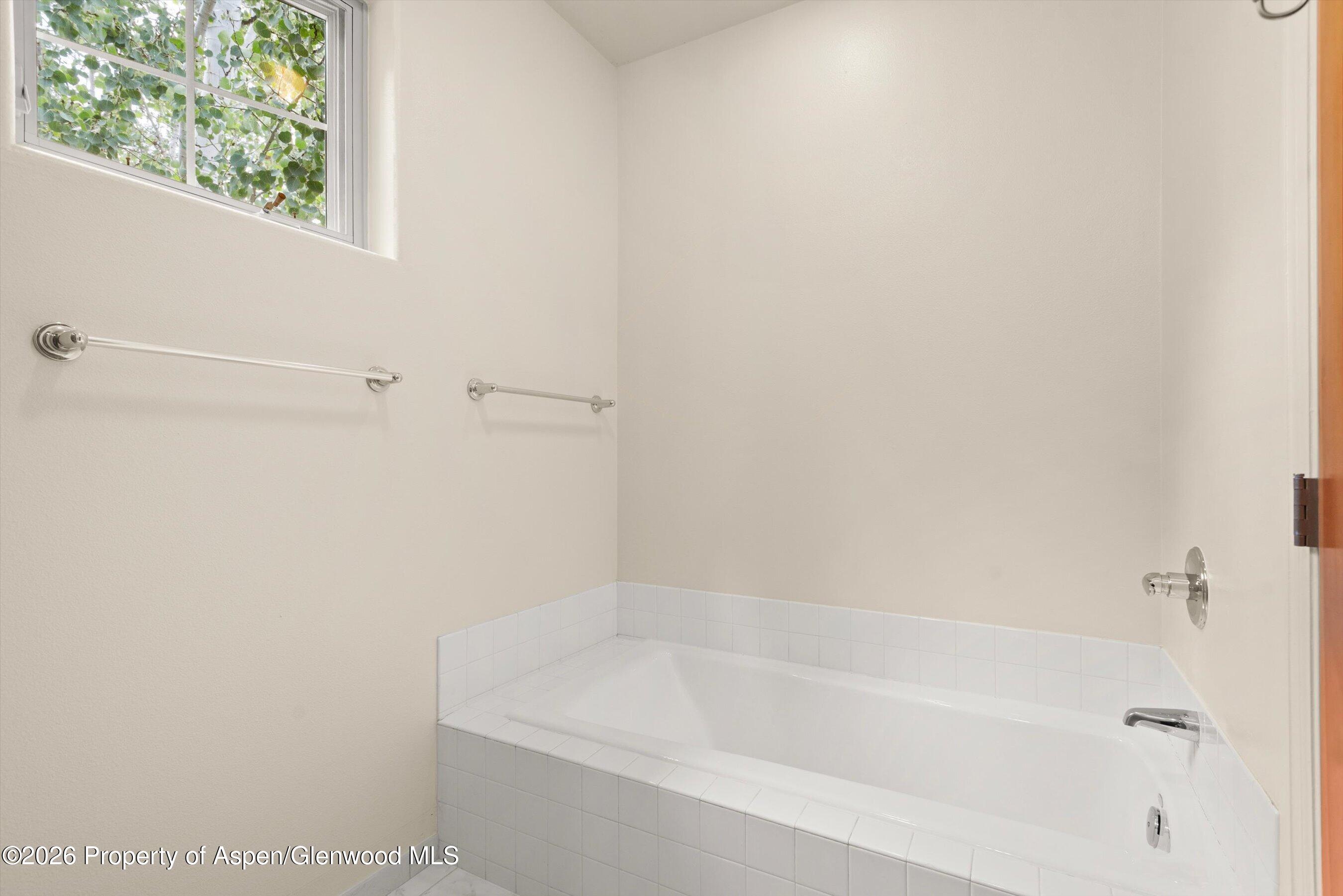 Bathroom with a white tiled bathtub beneath a high window overlooking leafy greenery.