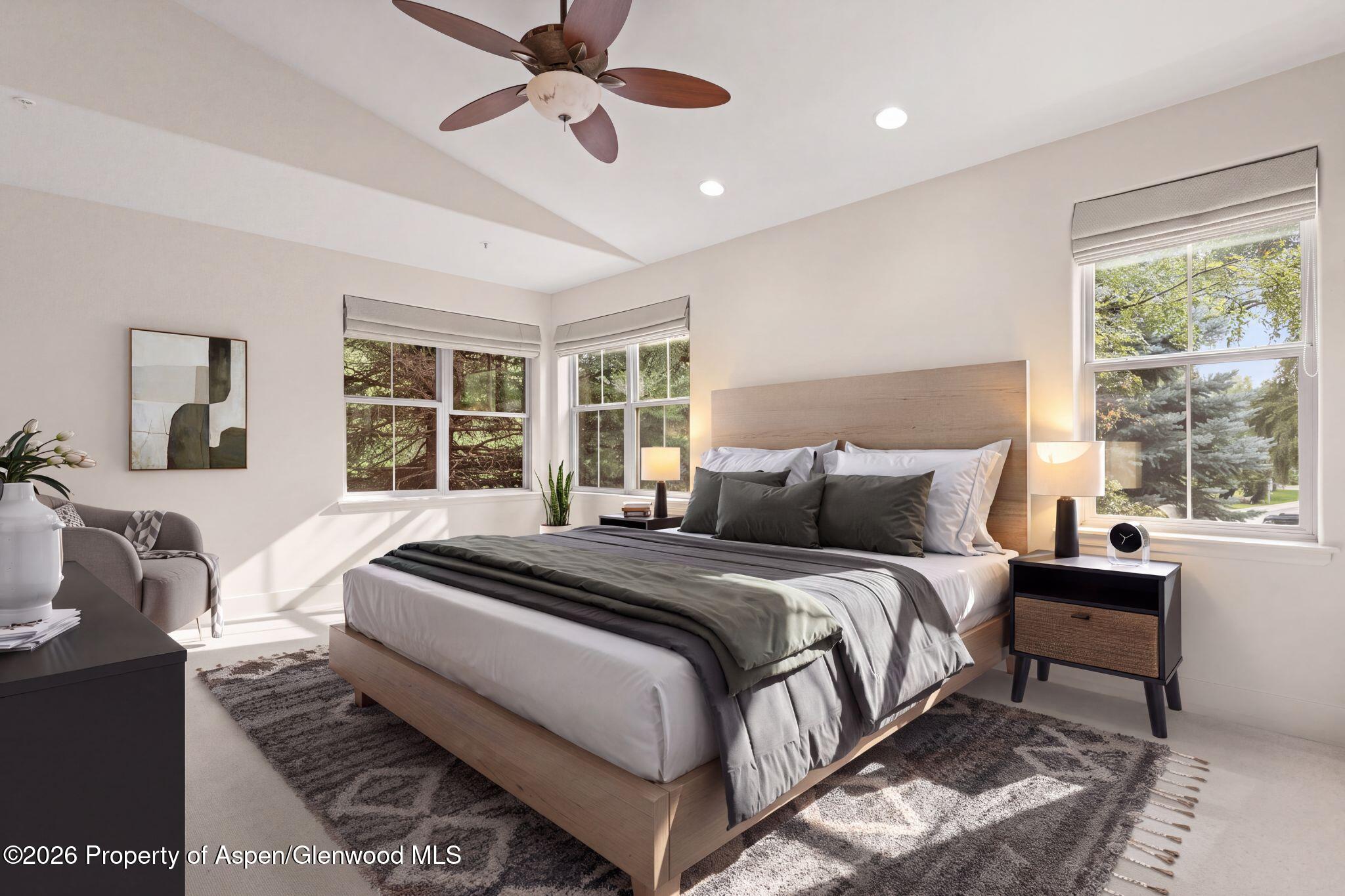 Bright bedroom with a wood-frame bed, gray bedding, ceiling fan, and windows overlooking trees.