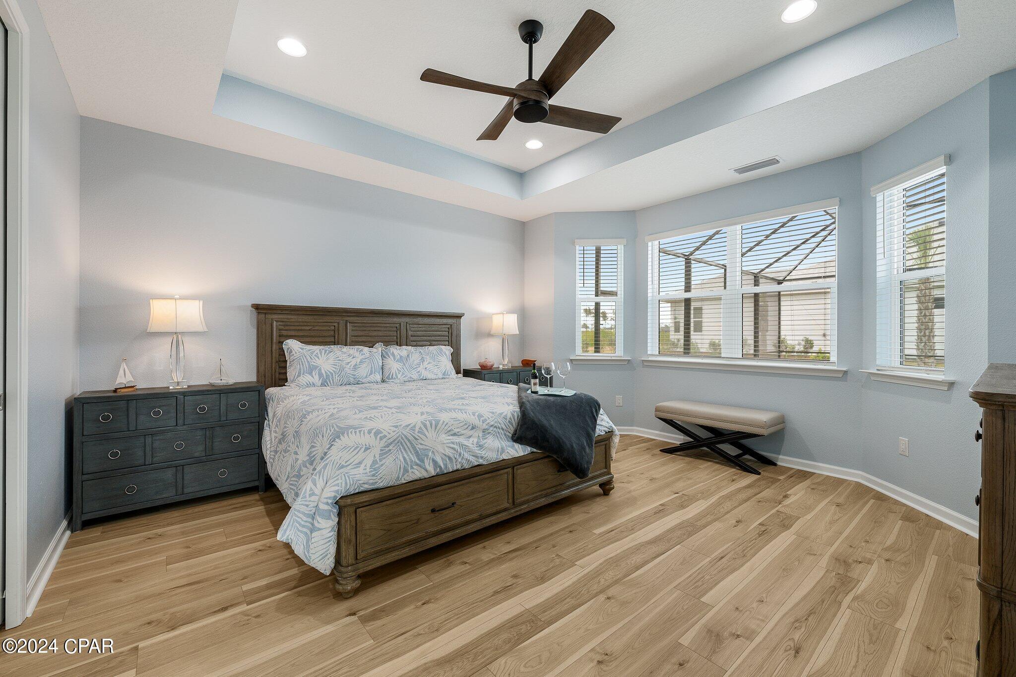 Furnished bedroom with a dark wood bed, pale blue walls, wide windows, and a tray ceiling fan.