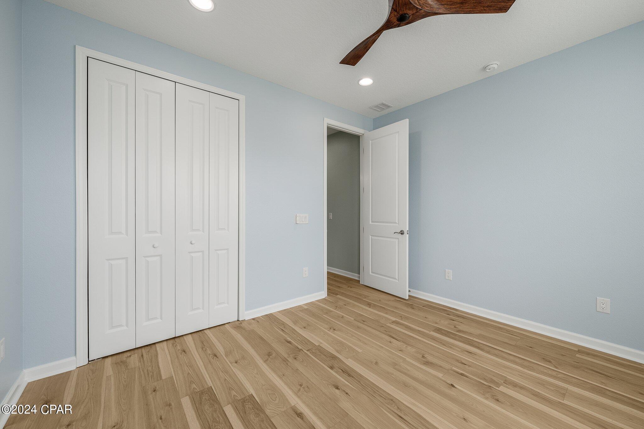 Empty bedroom with blue walls, white closet doors, wood-look flooring, and a ceiling fan.