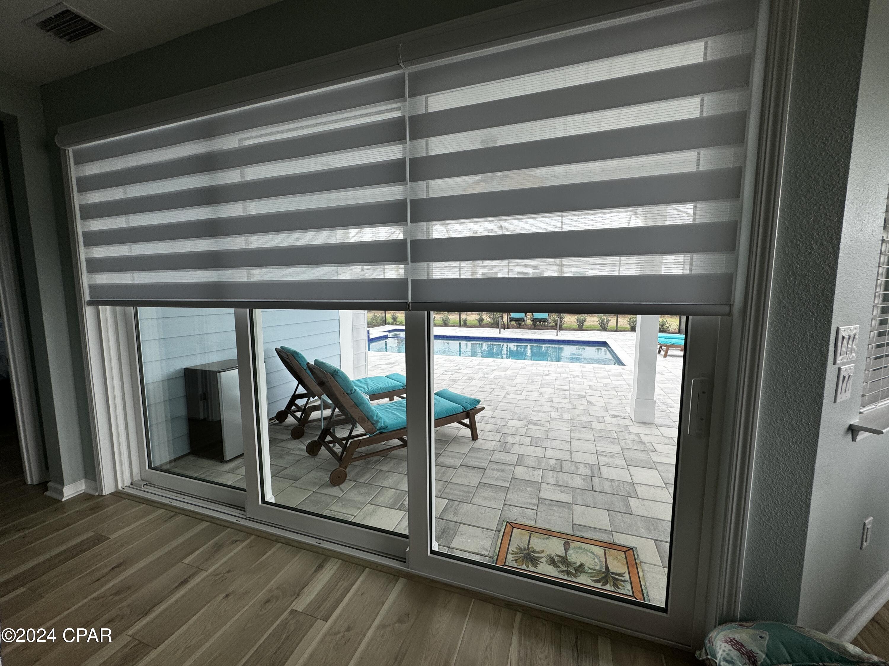 Sliding glass doors open to a paved patio with turquoise lounge chairs and a swimming pool.