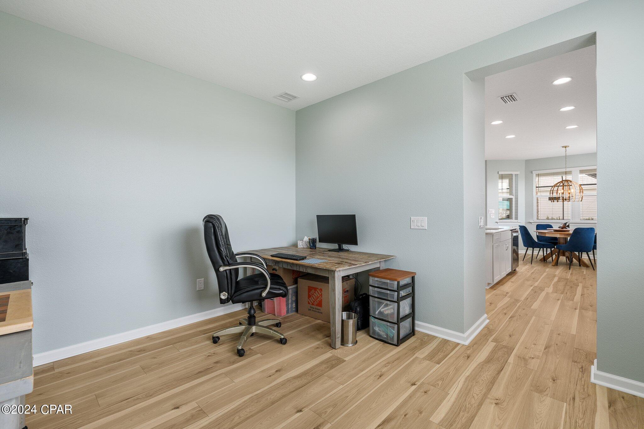 Home office with a rustic desk and black swivel chair, opening onto a dining area with blue chairs.
