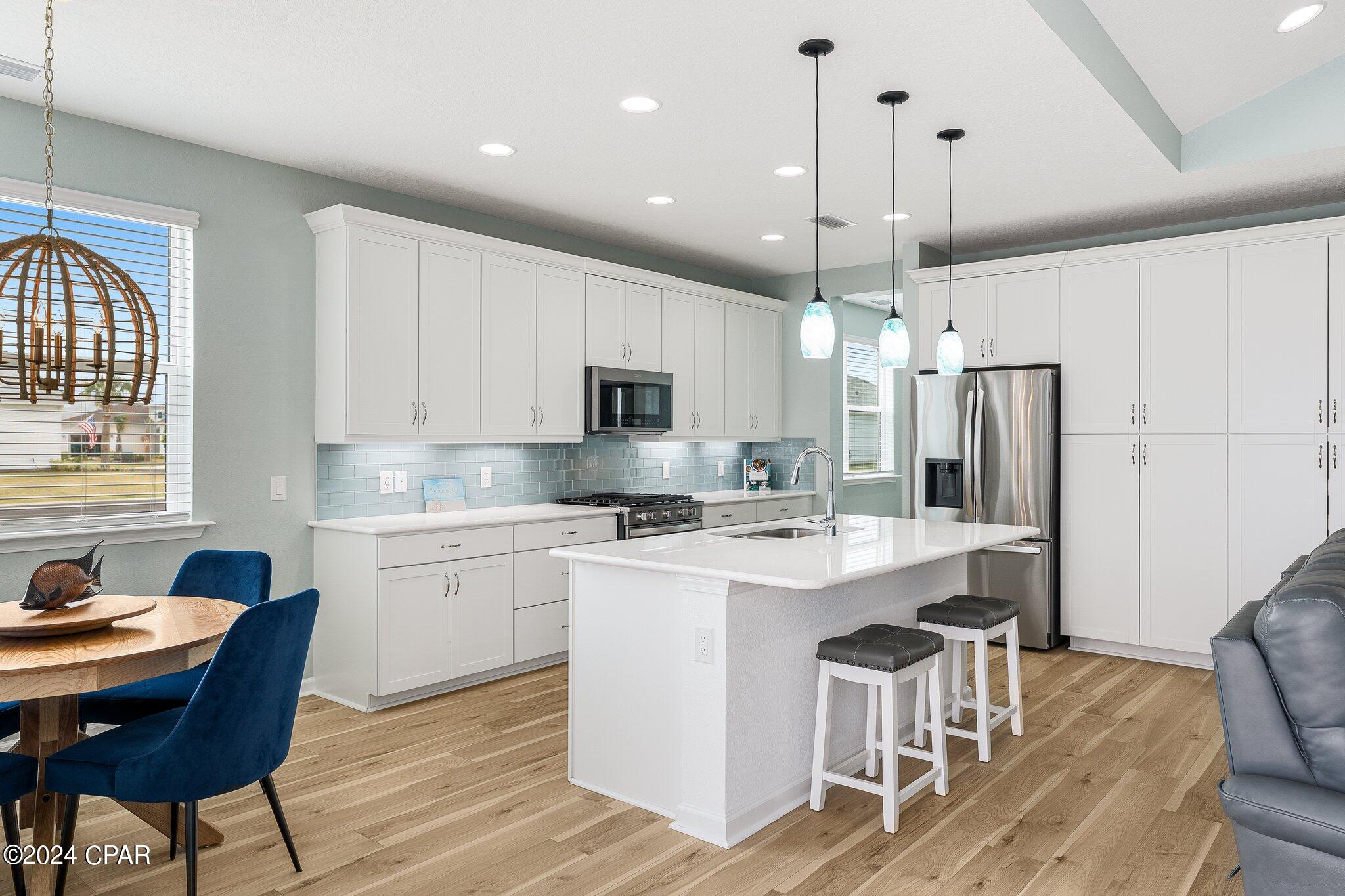 Open-plan kitchen with white cabinets, pale blue tile backsplash, stainless steel appliances, and a large island.