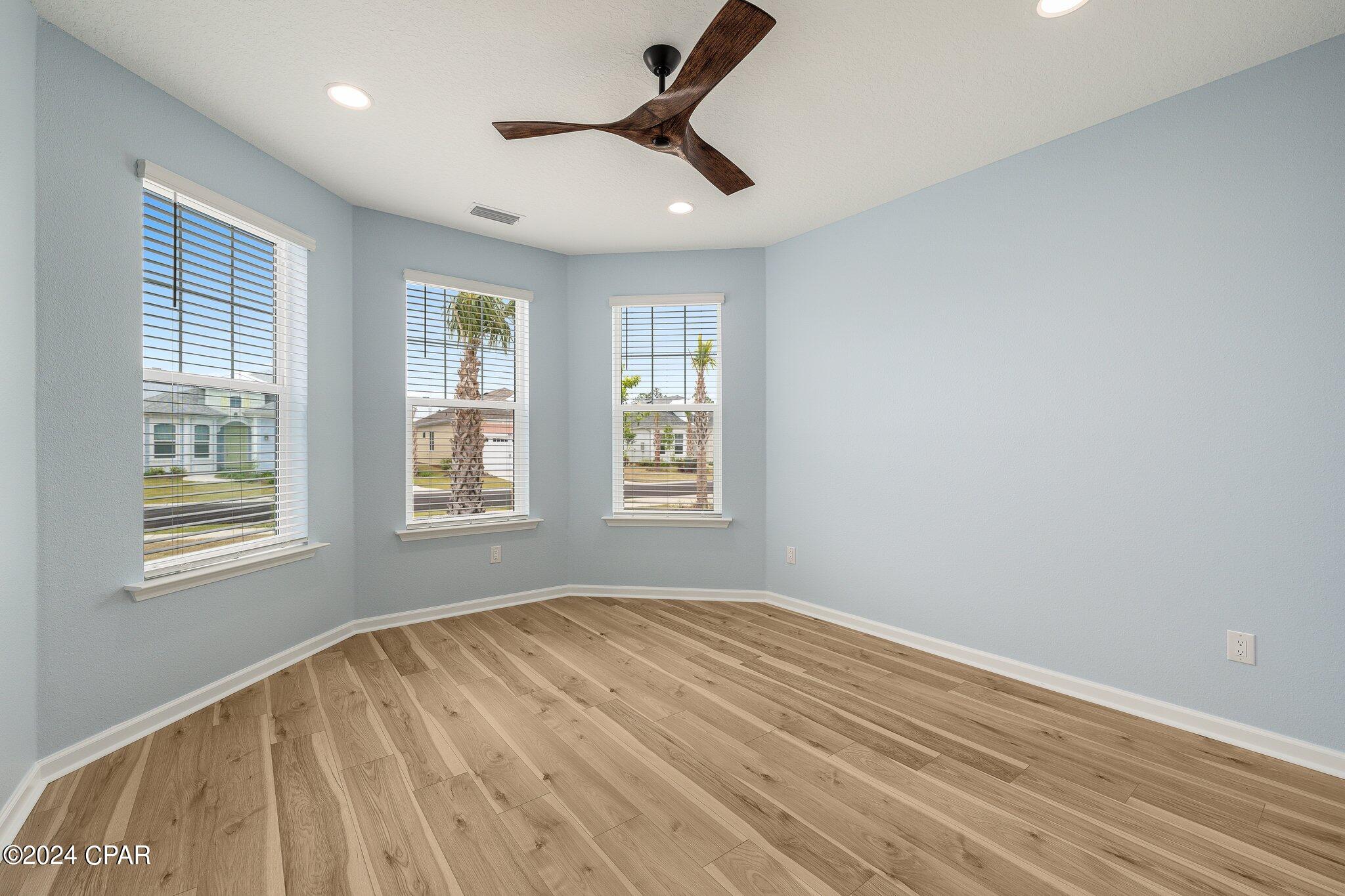 Vacant room with wood-look plank flooring, three windows, and a dark wood ceiling fan.