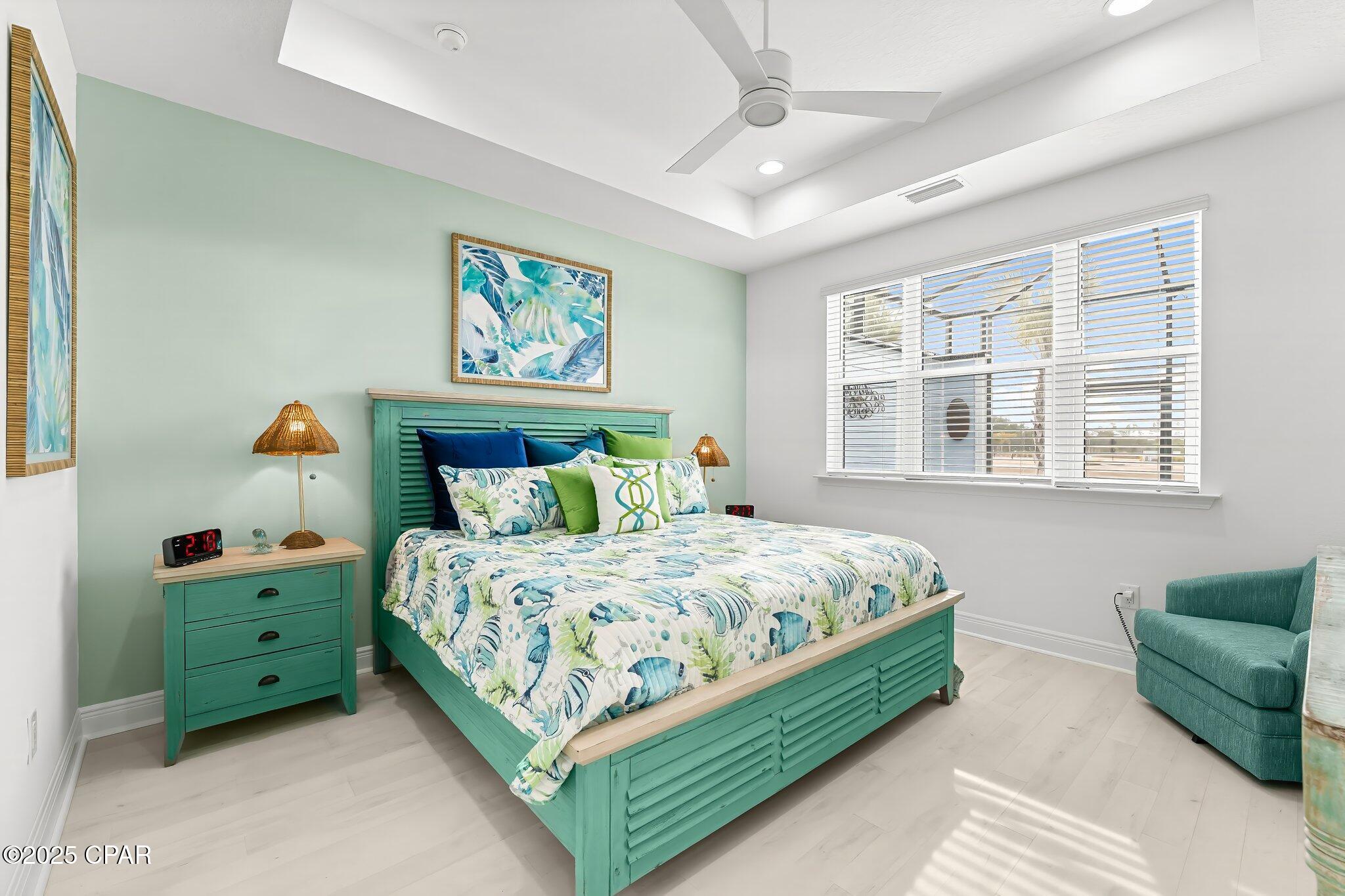 Furnished bedroom with a turquoise bed, tropical bedding, and large windows with white blinds.