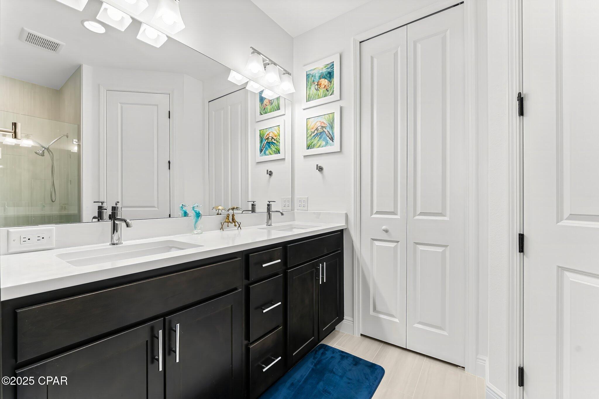 Bathroom with a double-sink vanity, dark cabinets, glass shower, and blue floor mat.