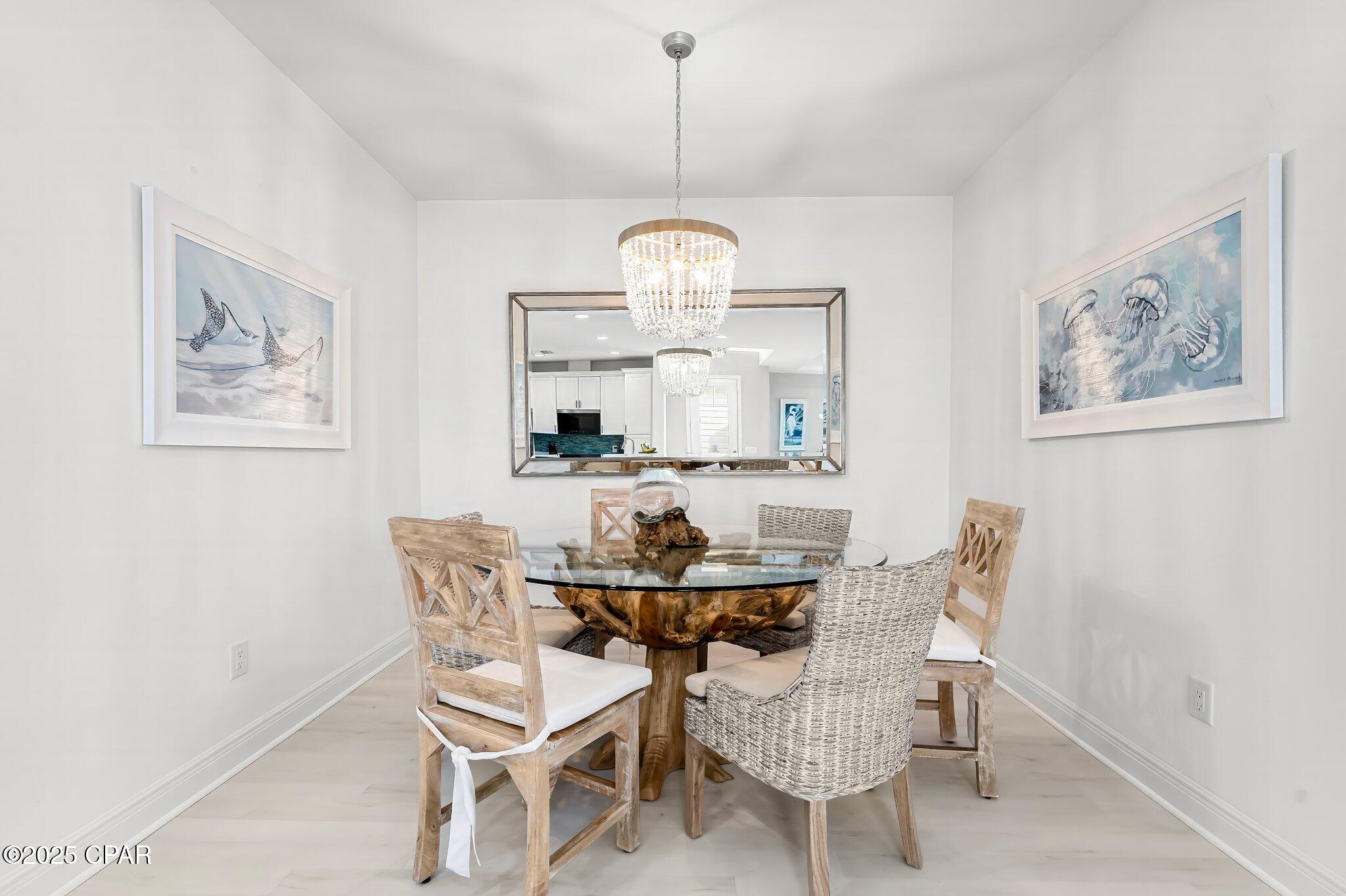 Furnished dining room with a glass-top table, woven chairs, crystal chandelier, and coastal artwork.